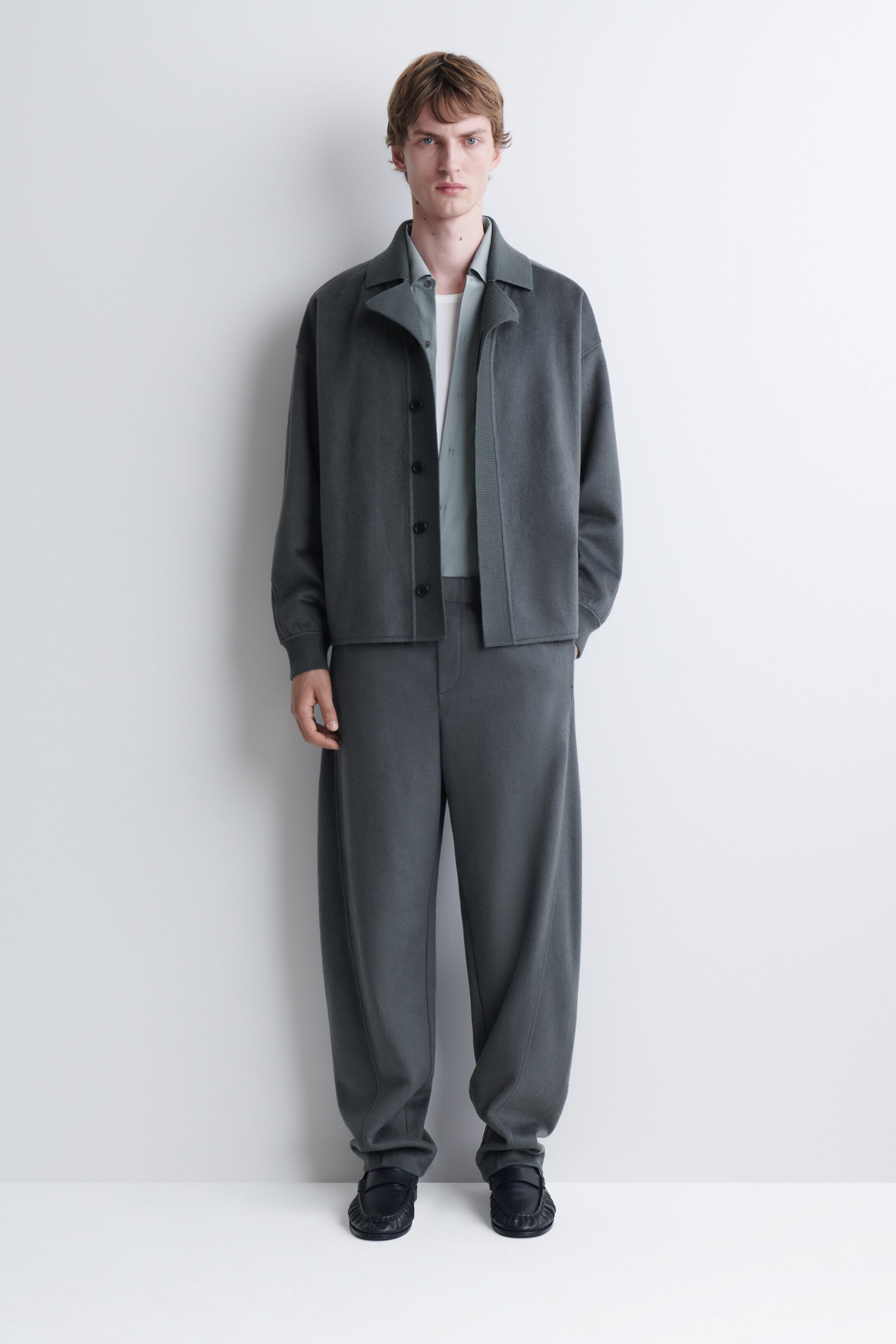 Le mannequin porte une veste en tricot gris foncé à col avec un pantalon ample assorti. La veste ouverte est superposée à une chemise boutonnée gris pâle et à un t-shirt blanc, le tout accompagné de mocassins en cuir noir. Le mannequin porte une veste en tricot gris foncé à col avec un pantalon ample assorti. La veste ouverte est superposée à une chemise boutonnée gris pâle et à un t-shirt blanc, le tout accompagné de mocassins en cuir noir.