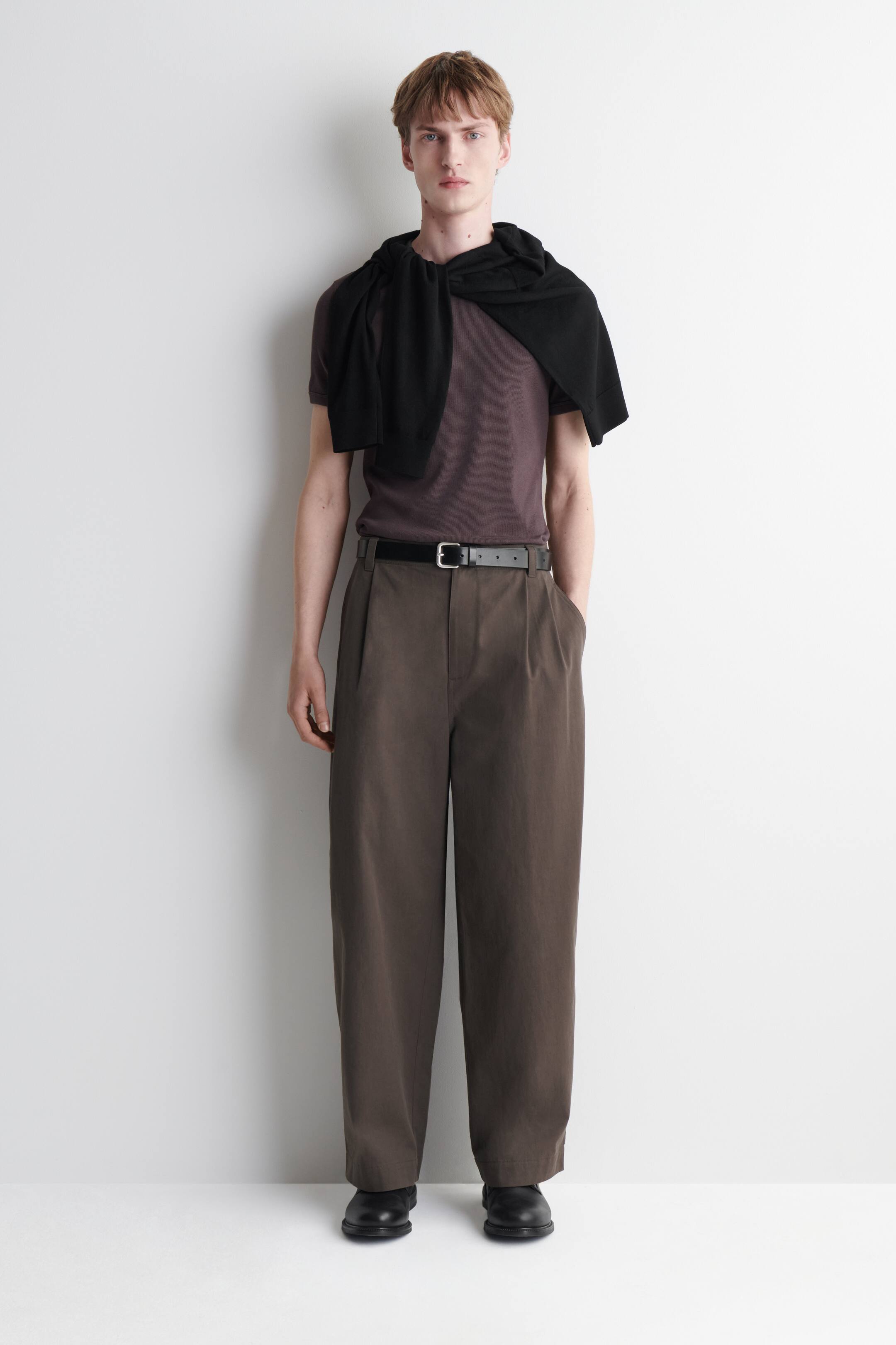 Größeres Bild ansehen: Ein Ganzkörper-Outfit wird aus einer Vorderansicht präsentiert, mit einem dunkelpflaumenfarbenen T-Shirt mit Rundhalsausschnitt, um dessen Schultern ein schwarzer Pullover gebunden ist. Das T-Shirt ist in eine dunkelolivfarbene, weit geschnittene, plissierte Hose gesteckt, die mit einem schwarzen Gürtel mit silberner Schnalle gehalten wird, und dazu werden schwarze Schuhe mit glatter Spitze getragen.