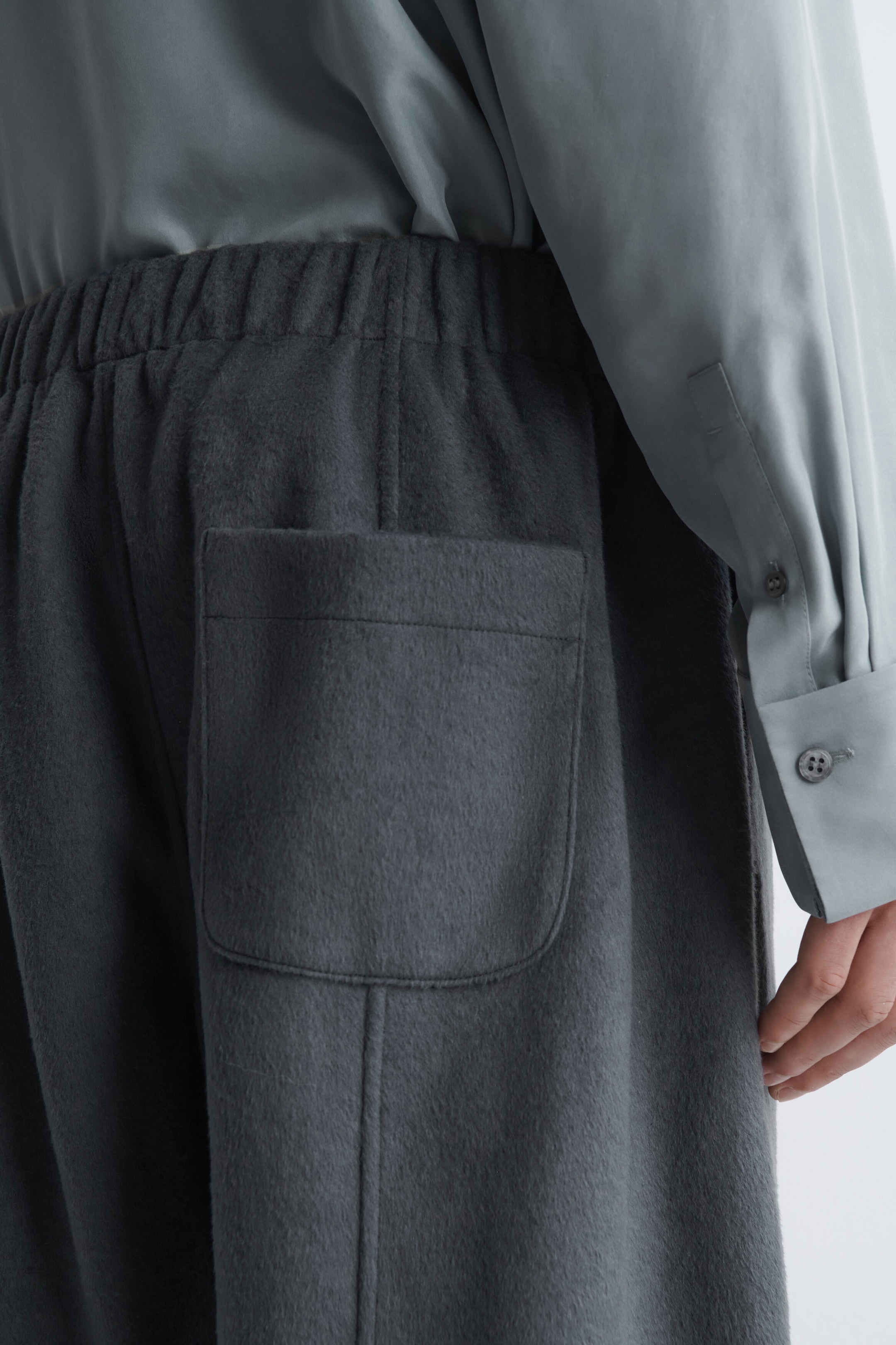 Le mannequin porte un pantalon en tissu texturé gris foncé, doté d’une taille élastiquée et d’une poche plaquée au dos. Le pantalon est agencé avec une chemise boutonnée vert sauge pâle rentrée à l’intérieur.