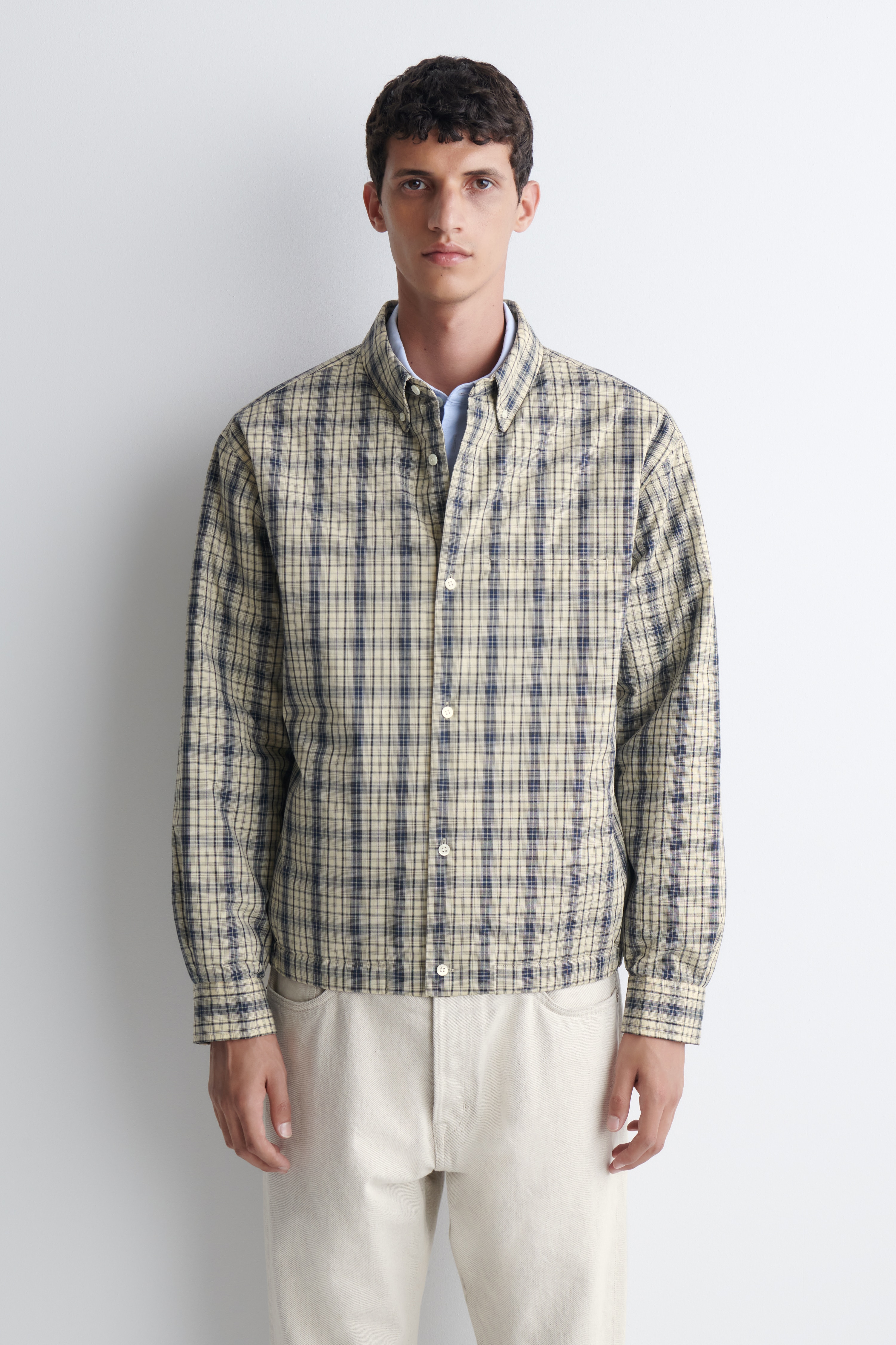 Vatovaná károvaná bavlnená overshirt je zobrazená spredu a prezentuje béžový a námornÃcky modrý kockovaný vzor s golierom na gombÃky a stredovým zapÃnanÃm na gombÃky. Overshirt má dlhé rukávy s manžetami na gombÃky, jednu náprsné vrecko a nariasený elastický spodný lem.