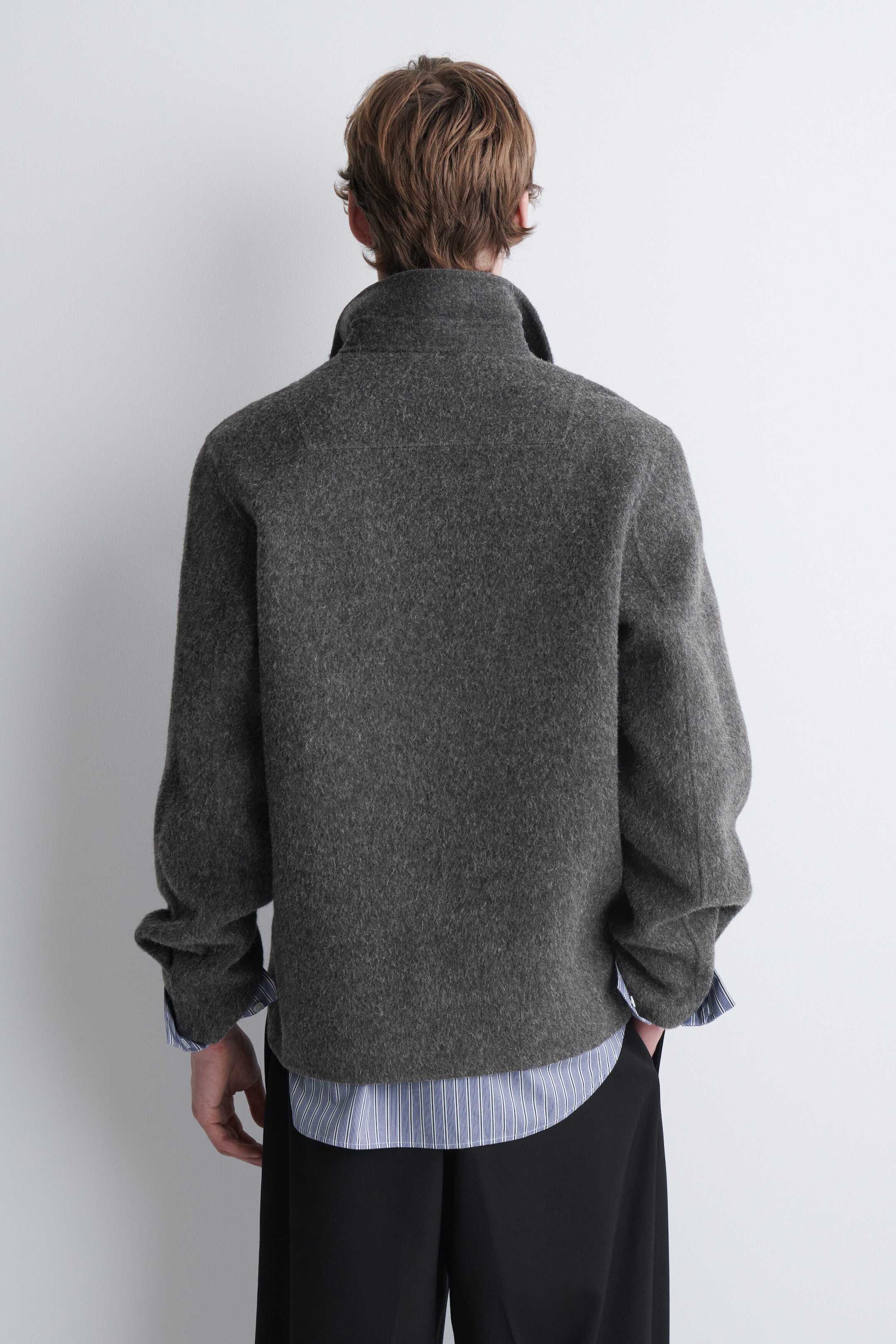 A gray textured wool jacket is being worn by the model, shown from the back. The jacket features a high collar and is layered over a blue-and-white striped shirt and black trousers.