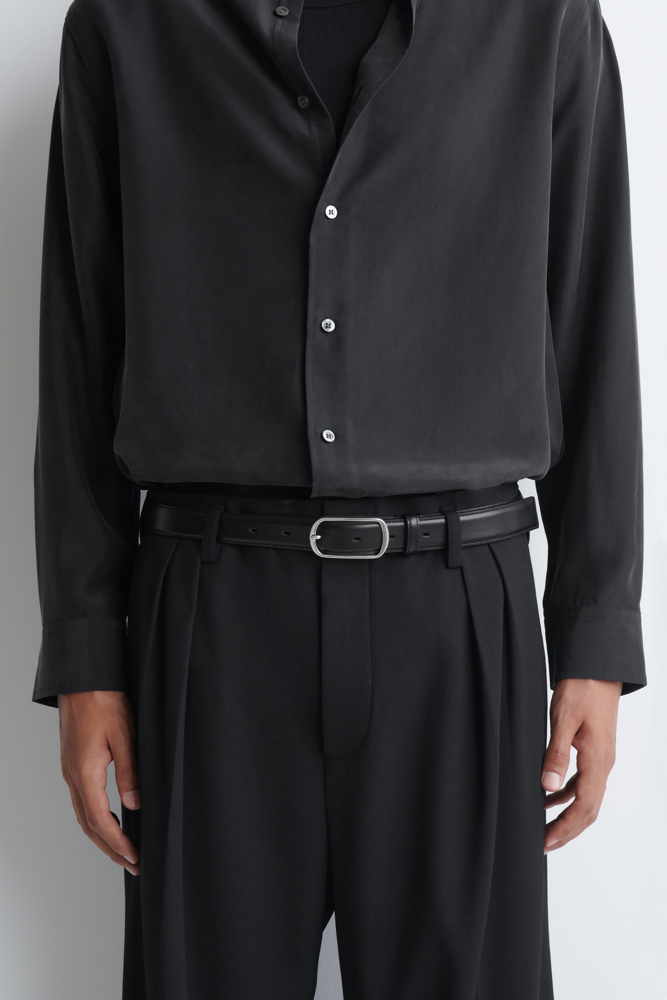 A black leather belt featuring a curved, silver-toned metal buckle is worn by the model through the belt loops of pleated black trousers. The belt secures the trousers and is styled with a tucked-in, long-sleeved black shirt. A black leather belt featuring a curved, silver-toned metal buckle is worn by the model through the belt loops of pleated black trousers. The belt secures the trousers and is styled with a tucked-in, long-sleeved black shirt.