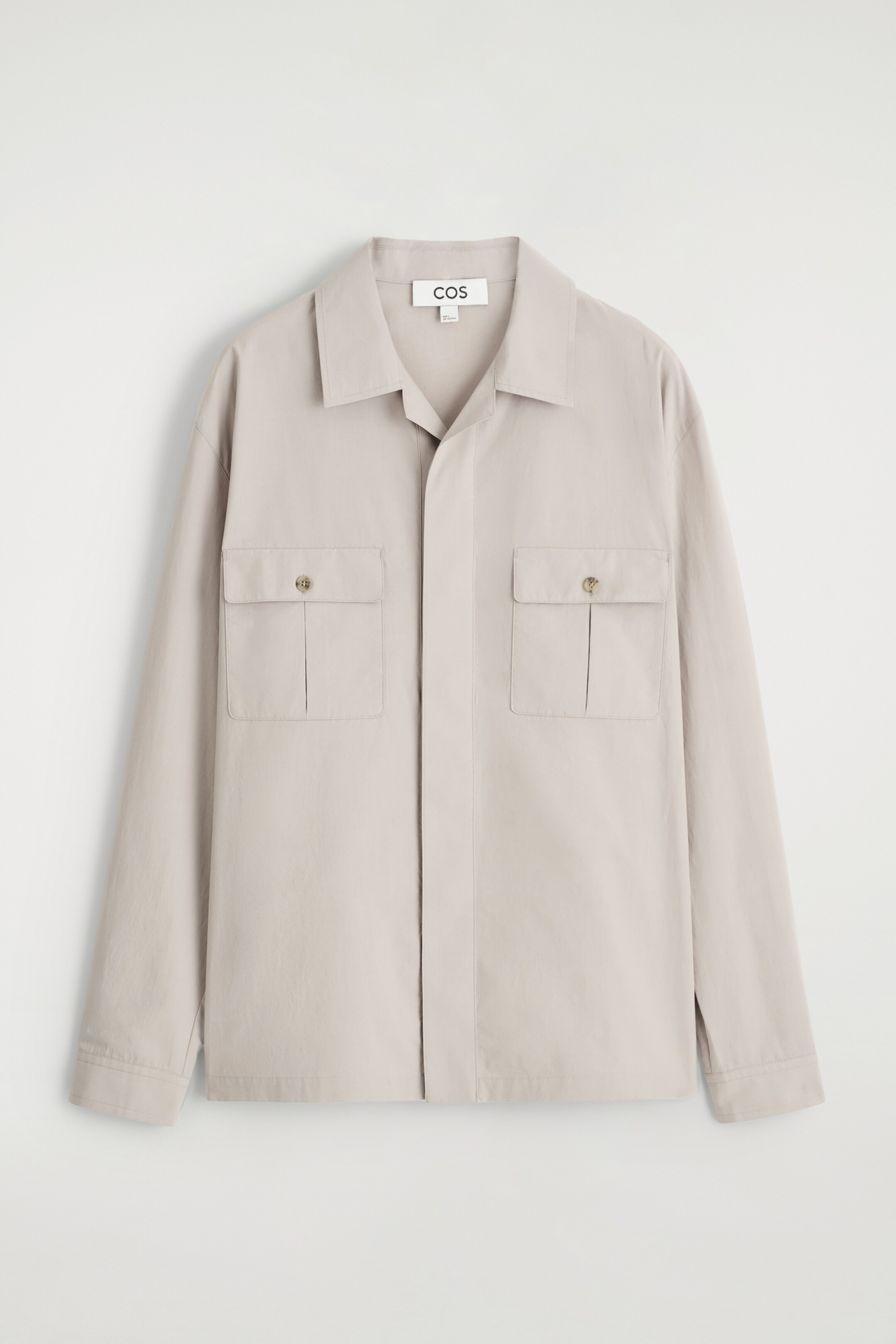 Größeres Bild anzeigen: Ein hellgraues Overshirt wird von vorne gezeigt, mit einer entspannten Passform, einem Umlegekragen und einer verdeckten Knopfleiste. Zwei große Klappentaschen sind auf der Brust sichtbar, jede mit einem runden Knopf gesichert.