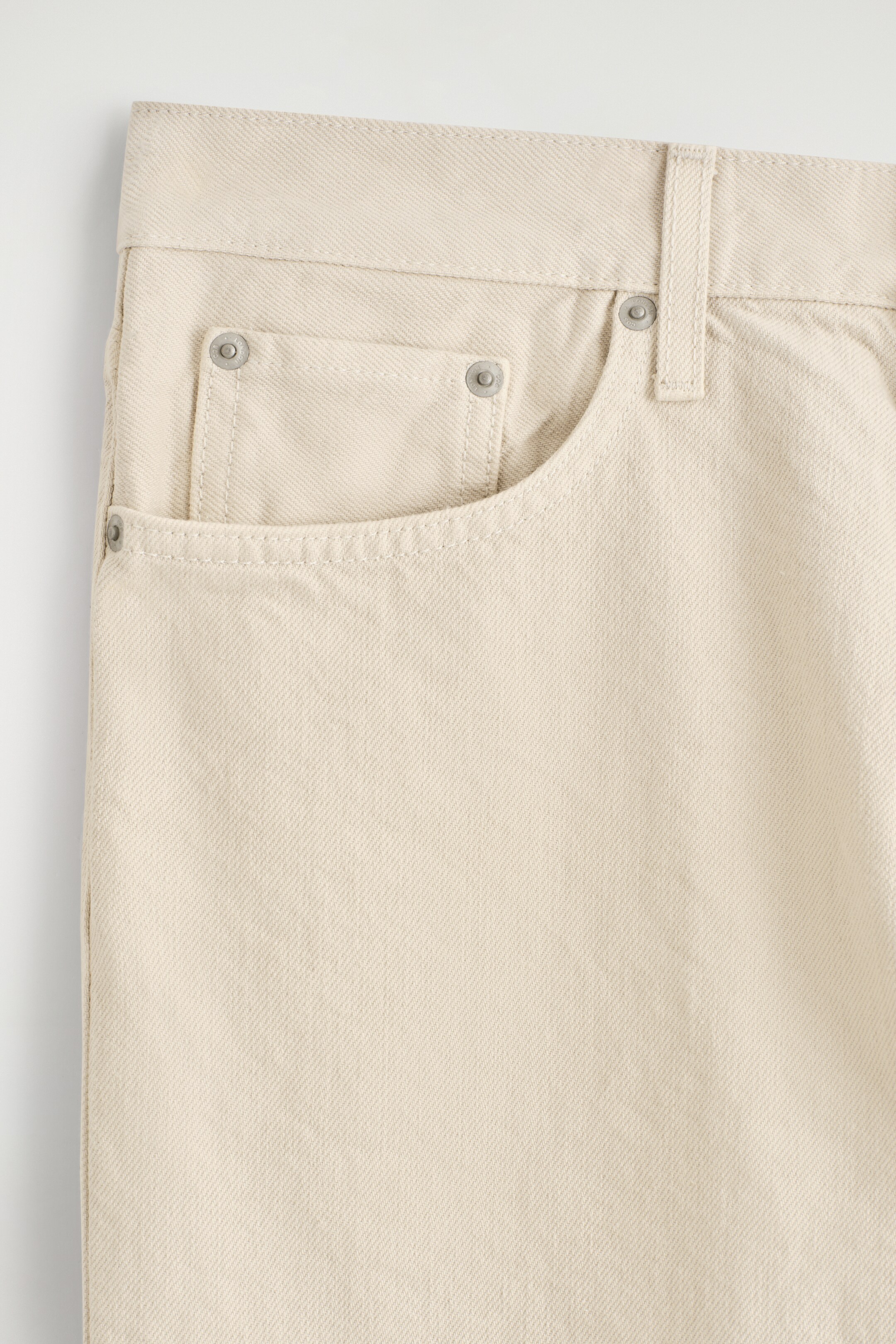 Größeres Bild anzeigen: Die Vorderseite der cremefarbenen Jeans zeigt auffällig ihre obere rechte Seite, mit einem Bund, einer Gürtelschlaufe, einer geschwungenen Vordertasche mit einer kleineren Münztasche und Metallnieten an den Taschenecken. Auffällige Steppnähte verzieren die Taschen und den Bund.