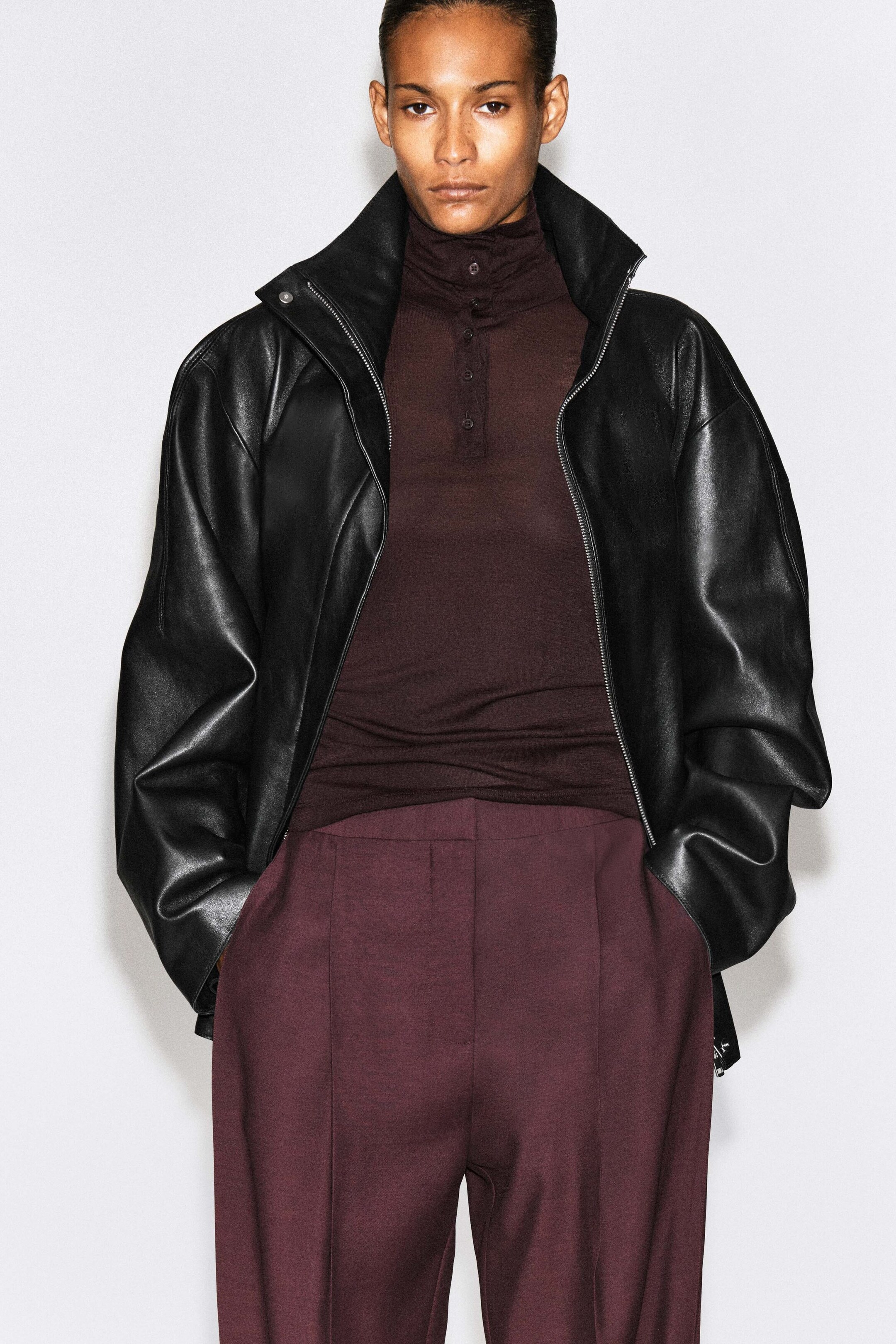 The model is wearing straight-leg pants in a deep burgundy hue, featuring structured front pleats. They are styled with a dark brown high-neck top and an open black leather jacket. The model is wearing straight-leg pants in a deep burgundy hue, featuring structured front pleats. They are styled with a dark brown high-neck top and an open black leather jacket.