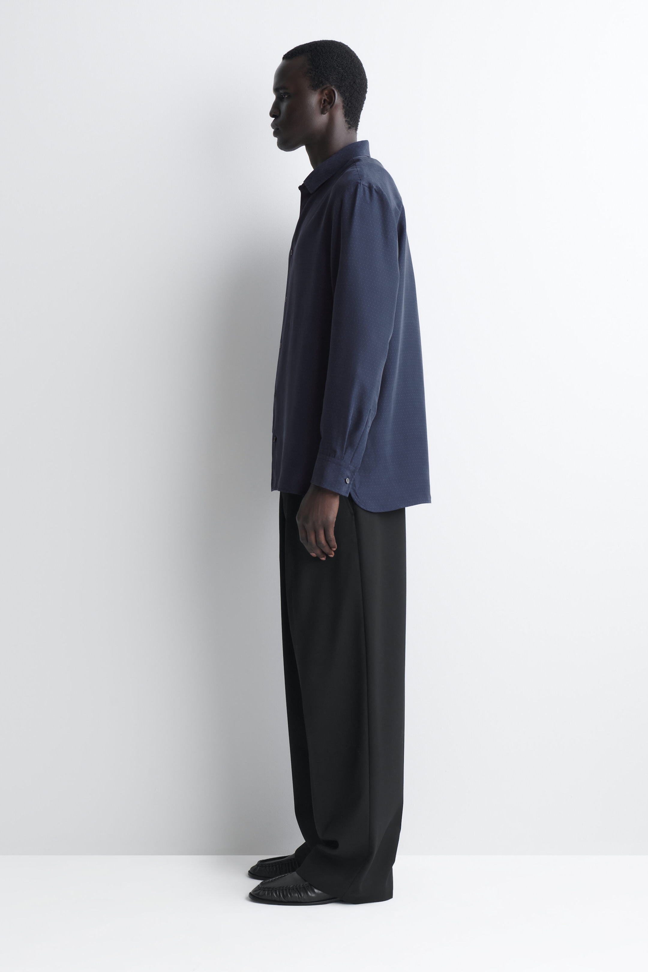 A navy blue sandwashed silk shirt is shown from the side, facing left, highlighting its relaxed fit, pointed collar, and curved hem. The long-sleeve shirt drapes loosely over wide-leg black trousers, showcasing its soft texture and buttoned cuffs. A navy blue sandwashed silk shirt is shown from the side, facing left, highlighting its relaxed fit, pointed collar, and curved hem. The long-sleeve shirt drapes loosely over wide-leg black trousers, showcasing its soft texture and buttoned cuffs.