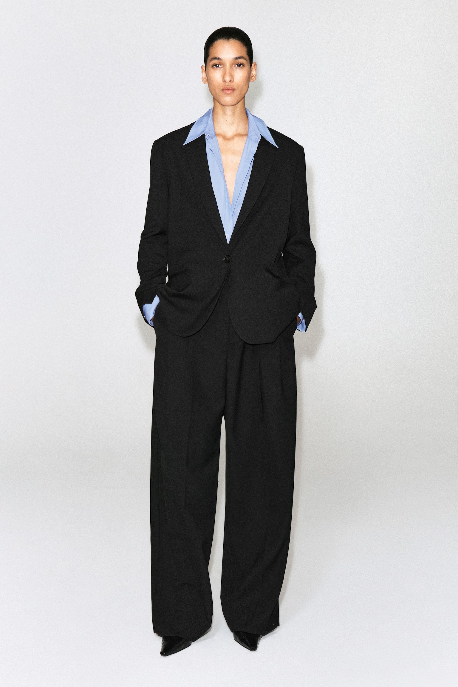 Un pantalon noir est présenté de FACE, doté d'une ceinture plate avec des passants de ceinture et de plis avant marqués. Des poches latérales en biais sont visibles sur chaque hanche, et le pantalon tombe en jambes larges et décontractées. - 1