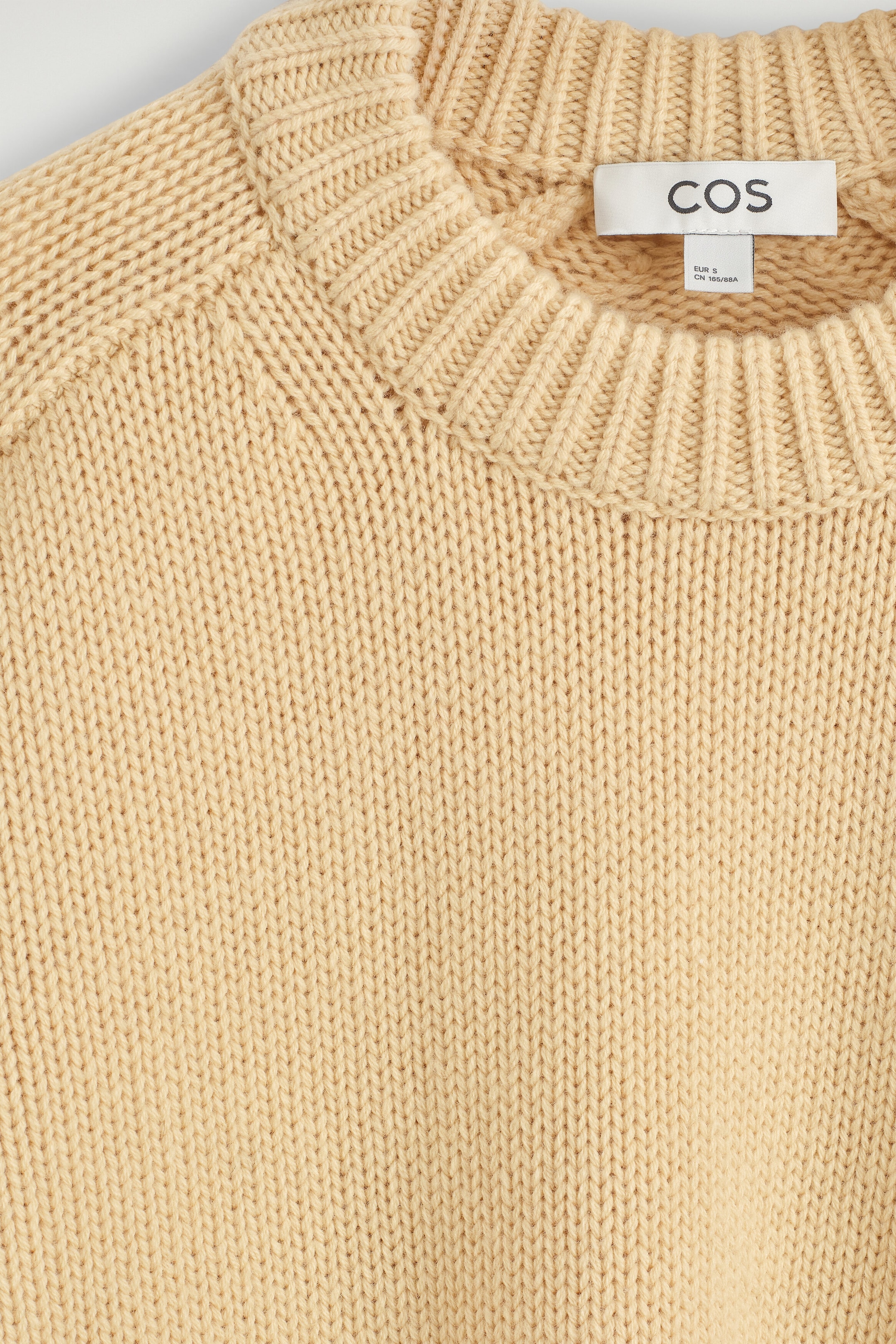 Ce chandail à col rond en laine beige, à la coupe arrondie, est présenté en gros plan de face, mettant en valeur son col rond côtelé épais et son motif de tricot vertical texturé. Le vêtement en tricot présente des détails d’épaules de style raglan ainsi qu’une étiquette de marque blanche cousue à l’intérieur de l’encolure.