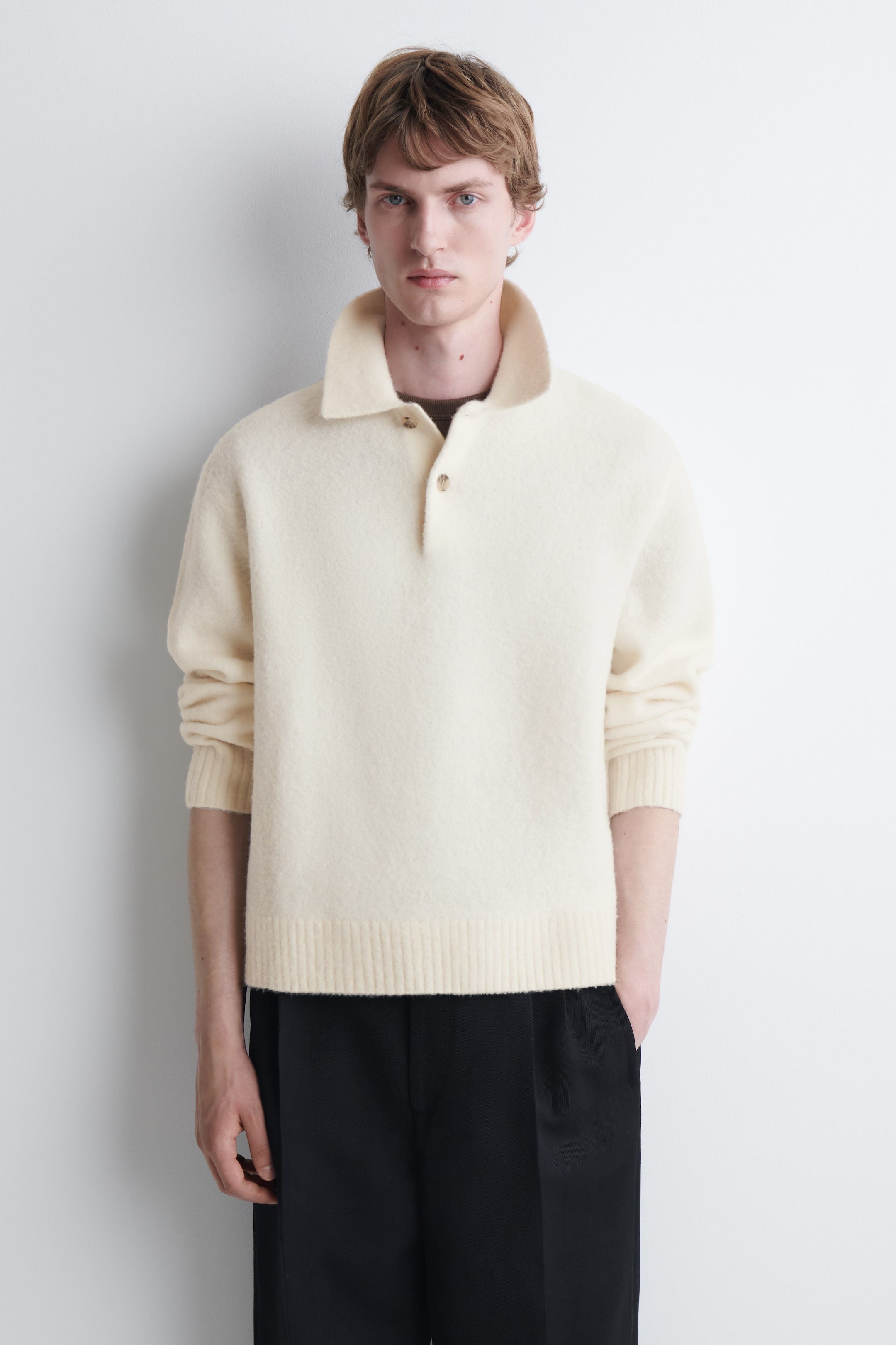 Visualizza l'immagine ingrandita: Il modello indossa una polo in lana spazzolata color crema, con colletto a punta, abbottonatura parziale e fondo a coste. La polo in maglia a maniche lunghe è indossata sopra un top scuro con scollo tondo ed è abbinata a pantaloni neri.