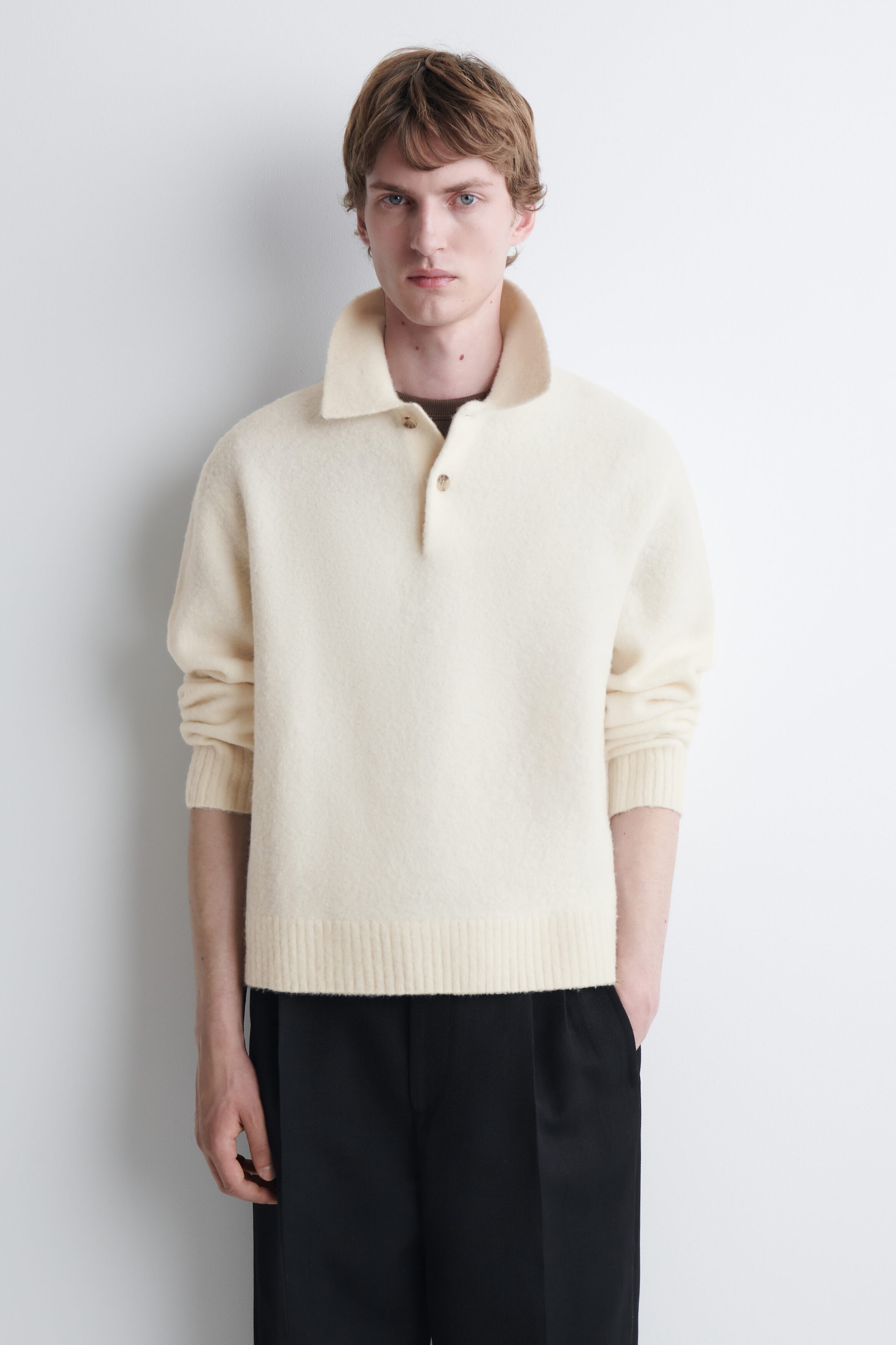 The model is wearing a cream-colored brushed wool polo shirt featuring a pointed collar, a partially buttoned placket, and a ribbed hem. The long-sleeve knit polo is layered over a dark crew-neck top and paired with black trousers.