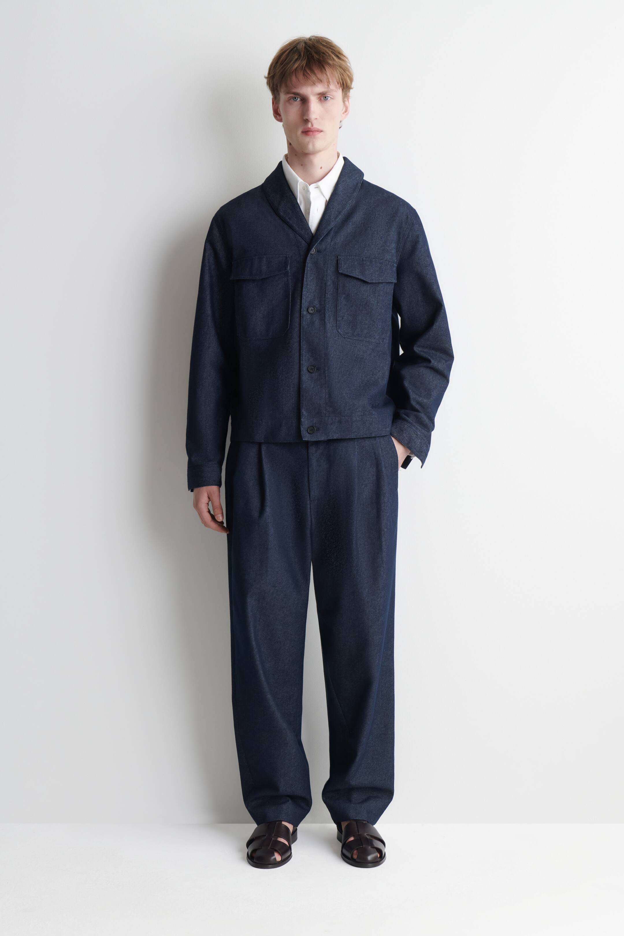 Größeres Bild anzeigen: Ein dunkelblaues Set im Denim-Look, bestehend aus einer passgenauen Jacke und einer weiten Hose, wird von vorne gezeigt. Die Jacke verfügt über einen Schalkragen, einen vorderen Drei-Knopf-Verschluss und zwei Brusttaschen mit Patten; darunter wird ein weißes Hemd mit Kragen getragen, während die weite Hose Frontfalten zeigt.