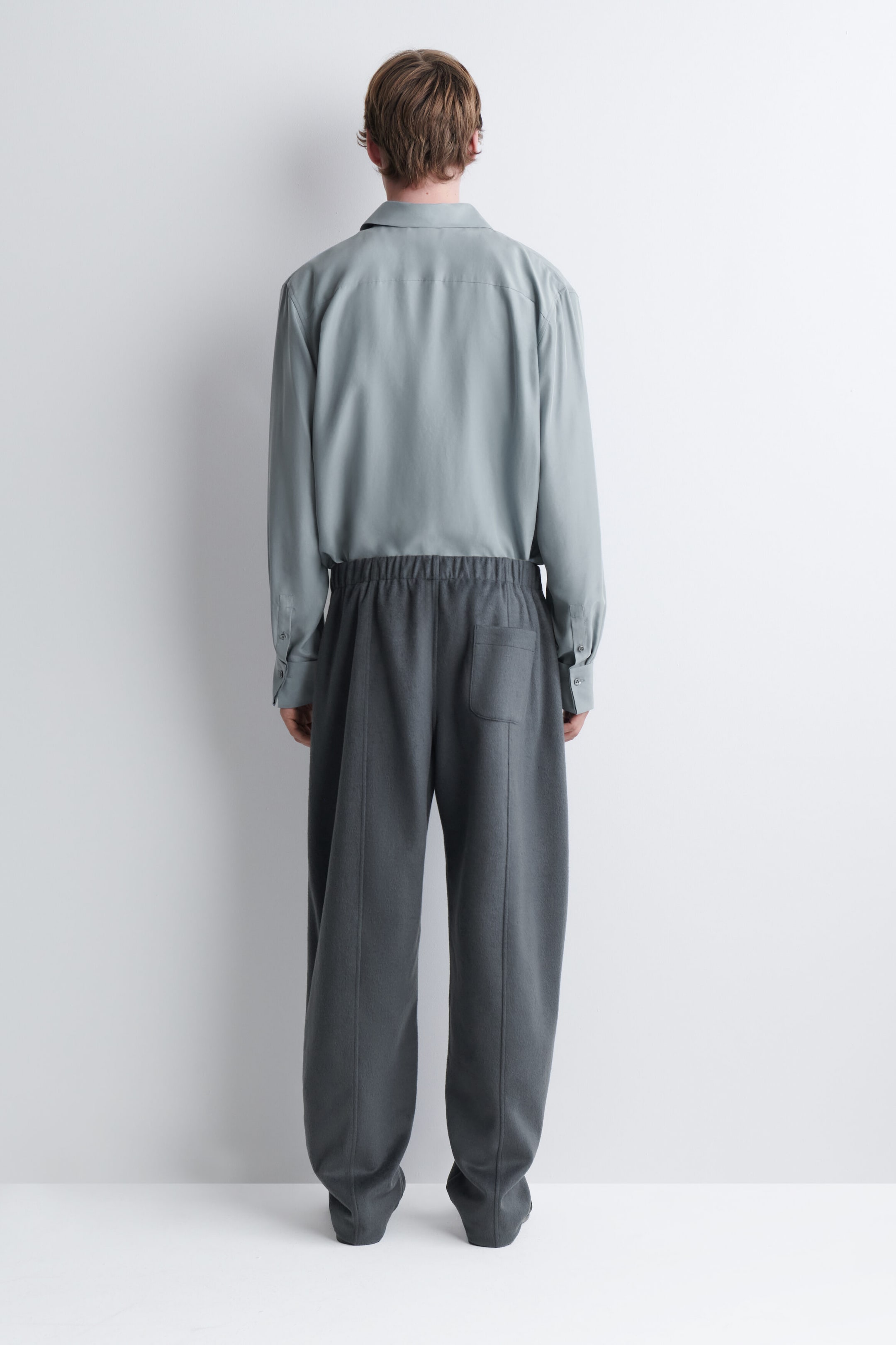 Un pantalon gris à jambe large, doté d’une taille élastique, de coutures centrales sur les jambes et d’une seule poche plaquée à l’arrière, est porté par le mannequin, vu de dos. Le pantalon est porté avec une chemise à manches longues vert sauge pâle à col, rentrée dans la taille.