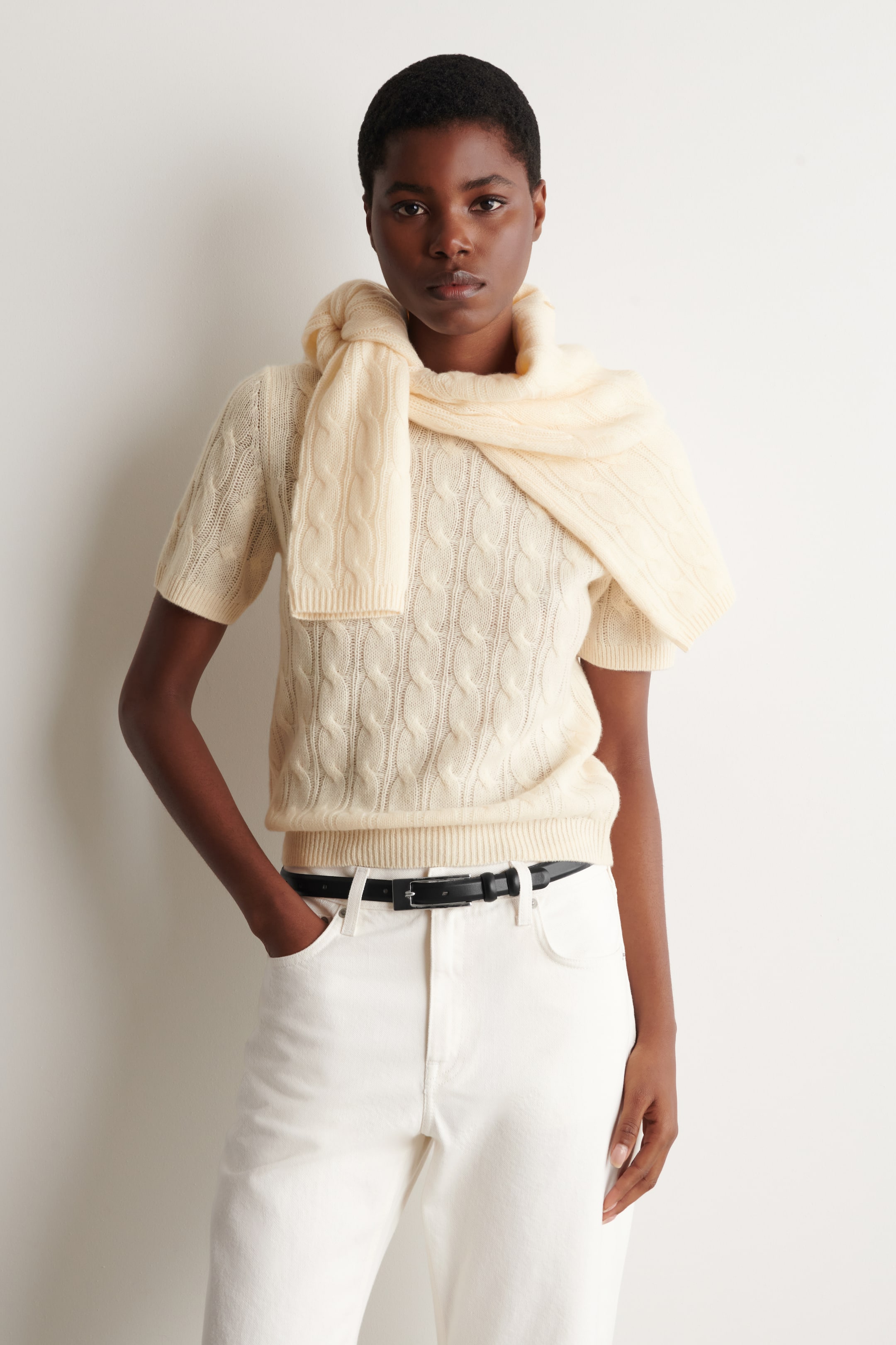 Agrandir l'image: Le mannequin porte un haut crème en maille torsadée à manches courtes, présentant un col rond, des manches courtes côtelées et un ourlet côtelé. Un pull assorti en maille torsadée crème est drapé sur les épaules du mannequin comme élément de styling.