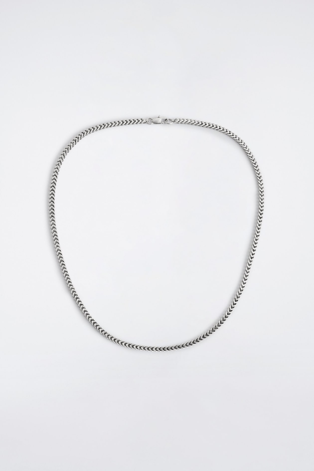 A silver-toned chain necklace is lying flat in a rounded shape, displaying a woven chevron-patterned design. The front-facing necklace features a lobster clasp closure at the top.