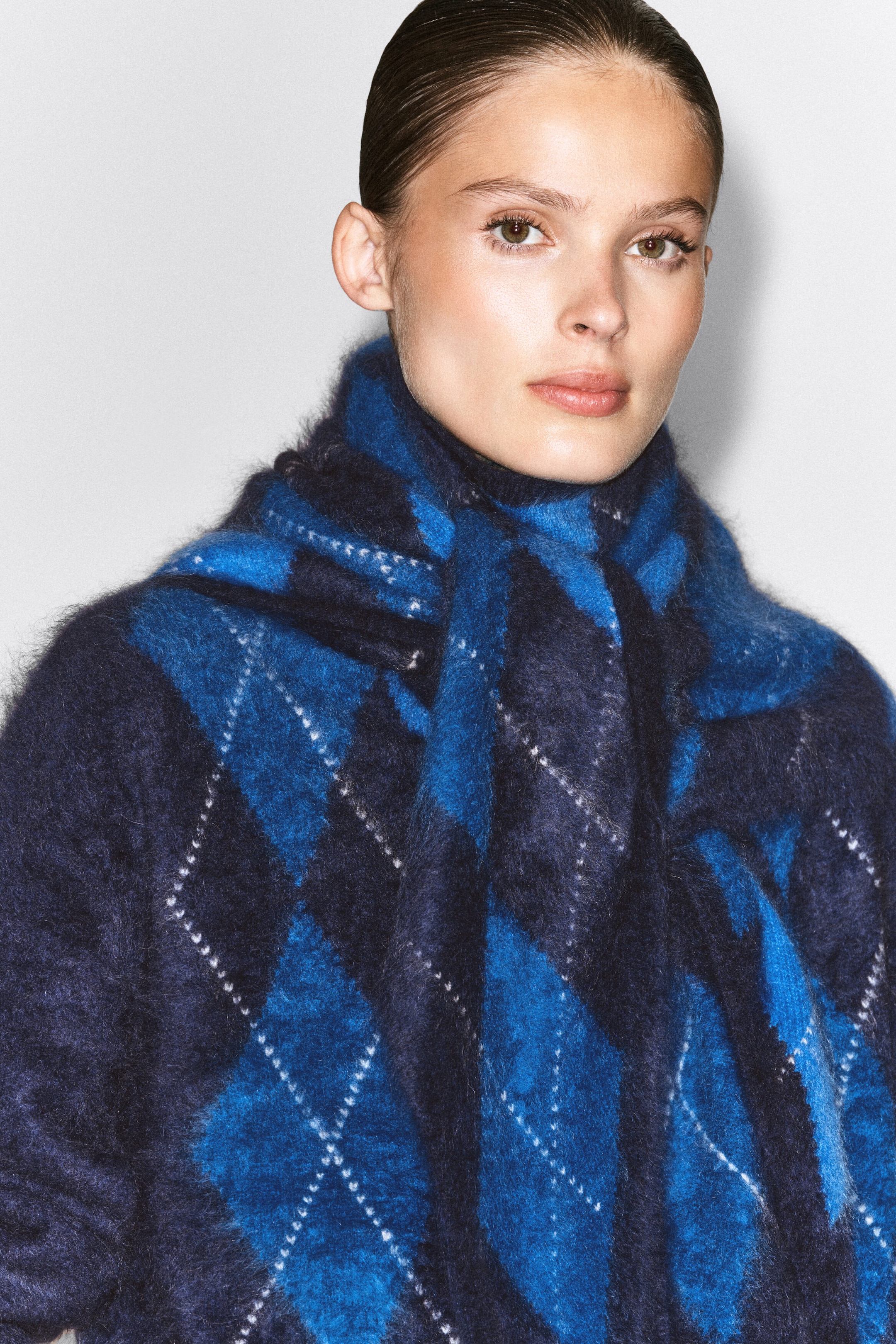 This argyle brushed pure cashmere funnel-neck jumper is worn by the model, featuring a fuzzy texture and a blue and navy diamond pattern. A matching argyle scarf is draped over the jumper around the model's neck and shoulders. This argyle brushed pure cashmere funnel-neck jumper is worn by the model, featuring a fuzzy texture and a blue and navy diamond pattern. A matching argyle scarf is draped over the jumper around the model's neck and shoulders.
