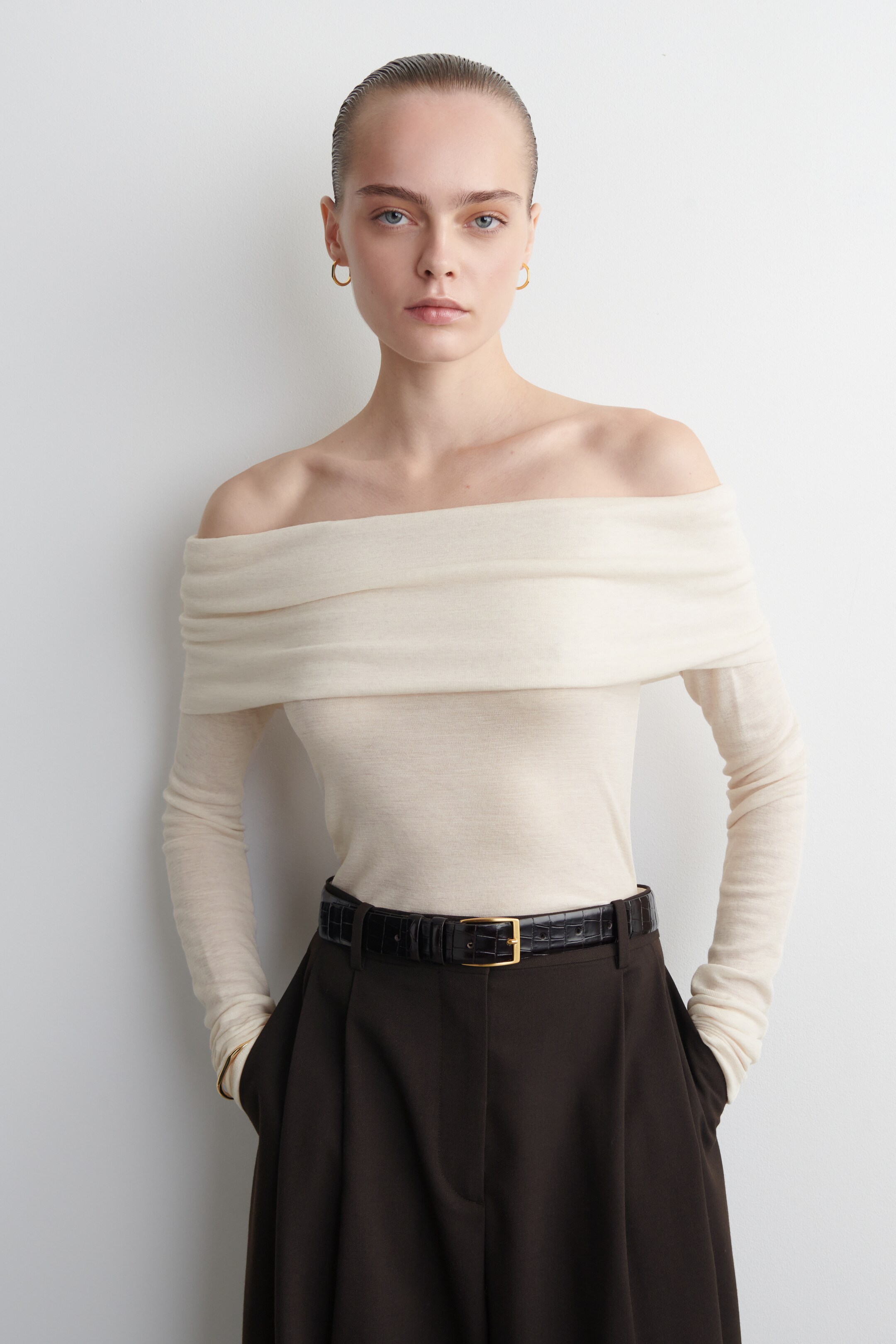 Agrandir l'image: Un haut beige clair à épaules dénudées est doté d'un large pli drapé sur le décolleté. Il est conçu avec de longues manches ajustées et se présente rentré dans un pantalon foncé et ceinturé avec une ceinture foncée, mettant en valeur sa silhouette ajustée.