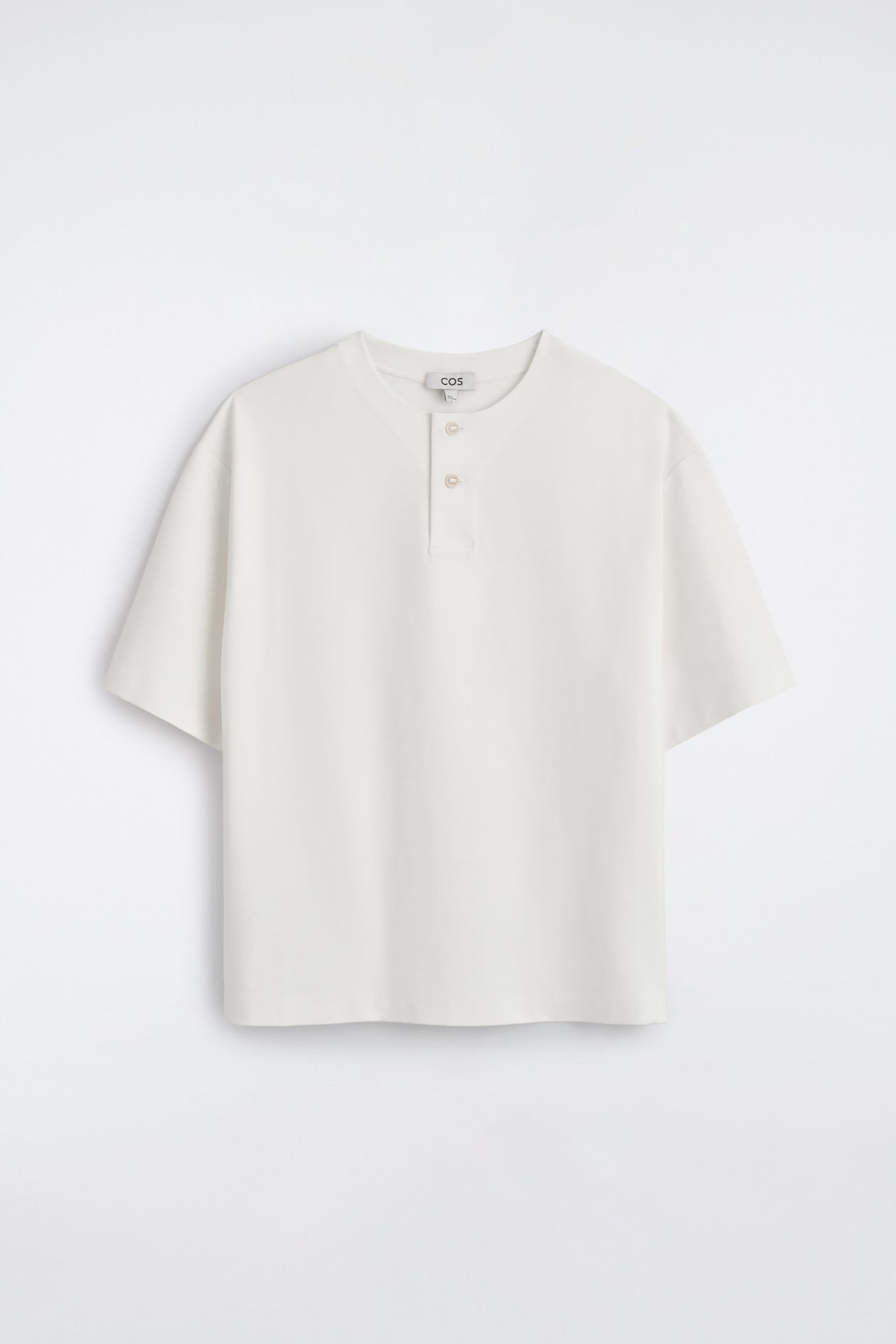 Un t-shirt henley oversize en jersey de coton blanc cassé est suspendu de face, affichant une coupe décontractée avec épaules tombantes et manches courtes larges. Il met en valeur un col rond avec patte de boutonnage partielle à deux boutons et un ourlet droit.