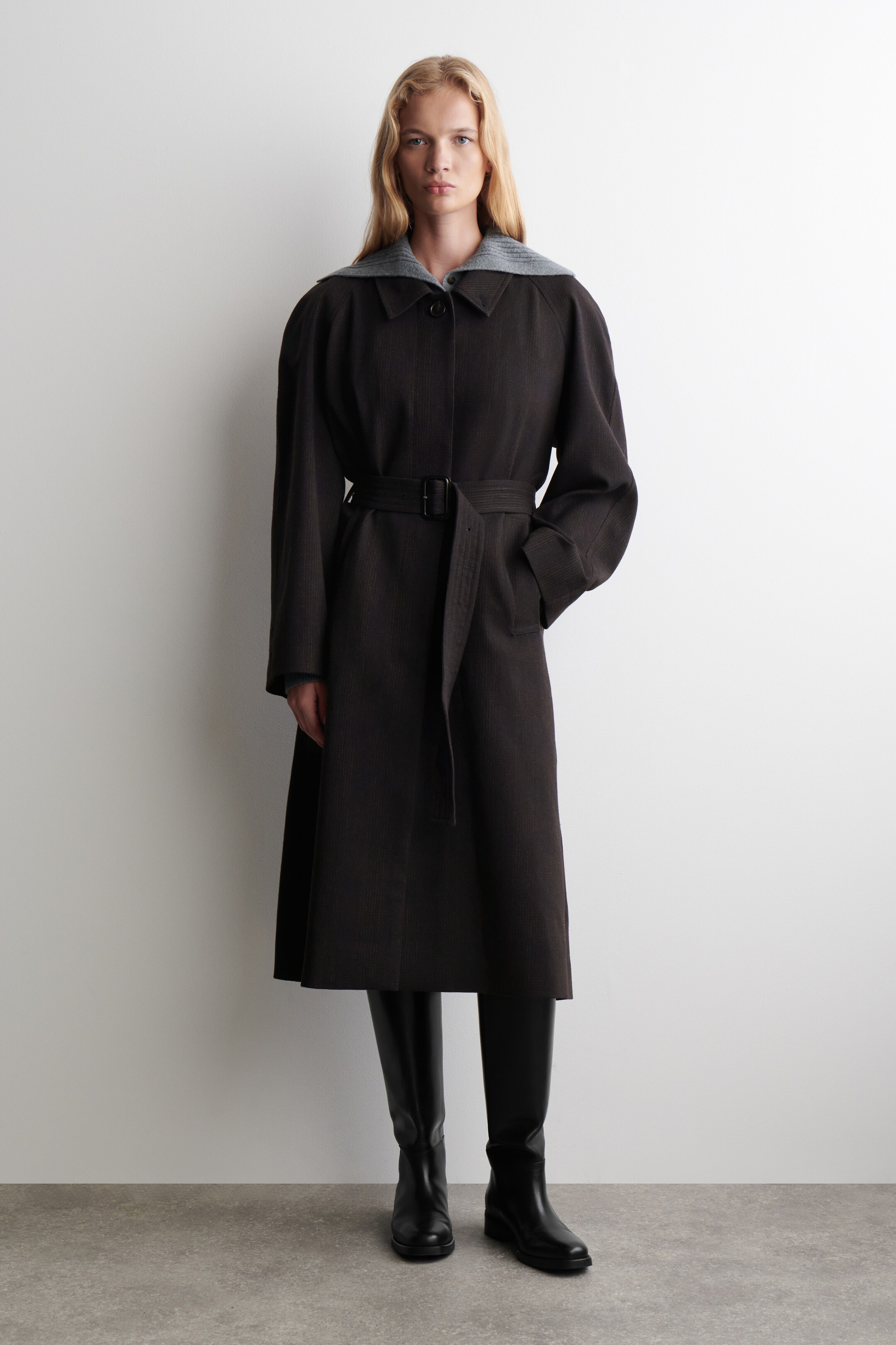 Agrandir l'image: Un manteau brun foncé, texturé, est présenté de face, s'étendant jusqu'à mi-mollet avec une silhouette surdimensionnée et des manches larges. Il est ceinturé à la taille par une ceinture en tissu assorti avec une boucle noire, et un vêtement en tricot côtelé gris est visible sous son col pointu.