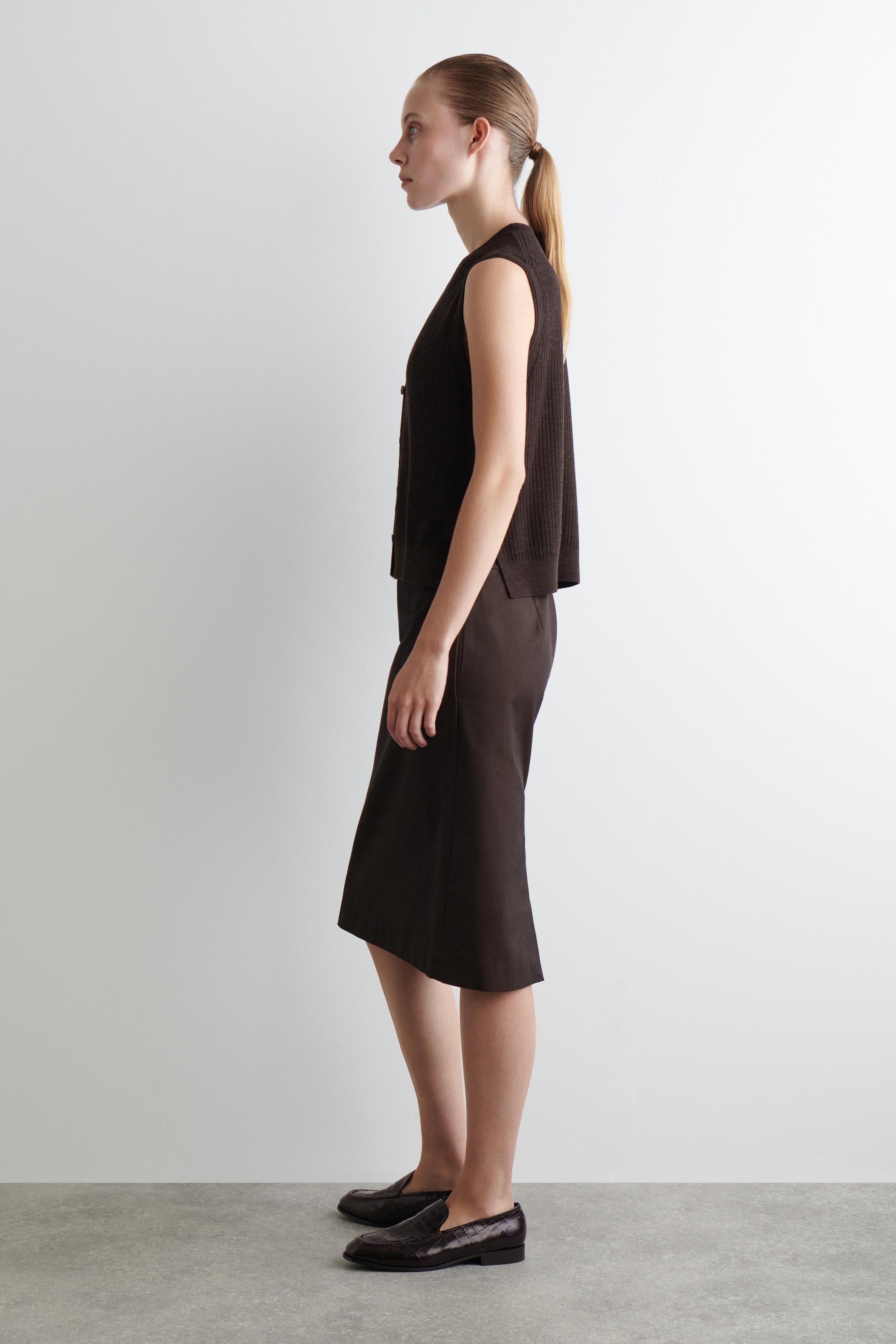 Ingrandisci l'immagine: Un ensemble a due pezzi di colore marrone scuro è esposto in un profilo laterale. Il top senza maniche in maglia a coste con chiusura a bottoni presenta una vestibilità rilassata, mentre la gonna midi coordinata mostra una silhouette dritta. Mocassini testurizzati marrone scuro completano il look coordinato.