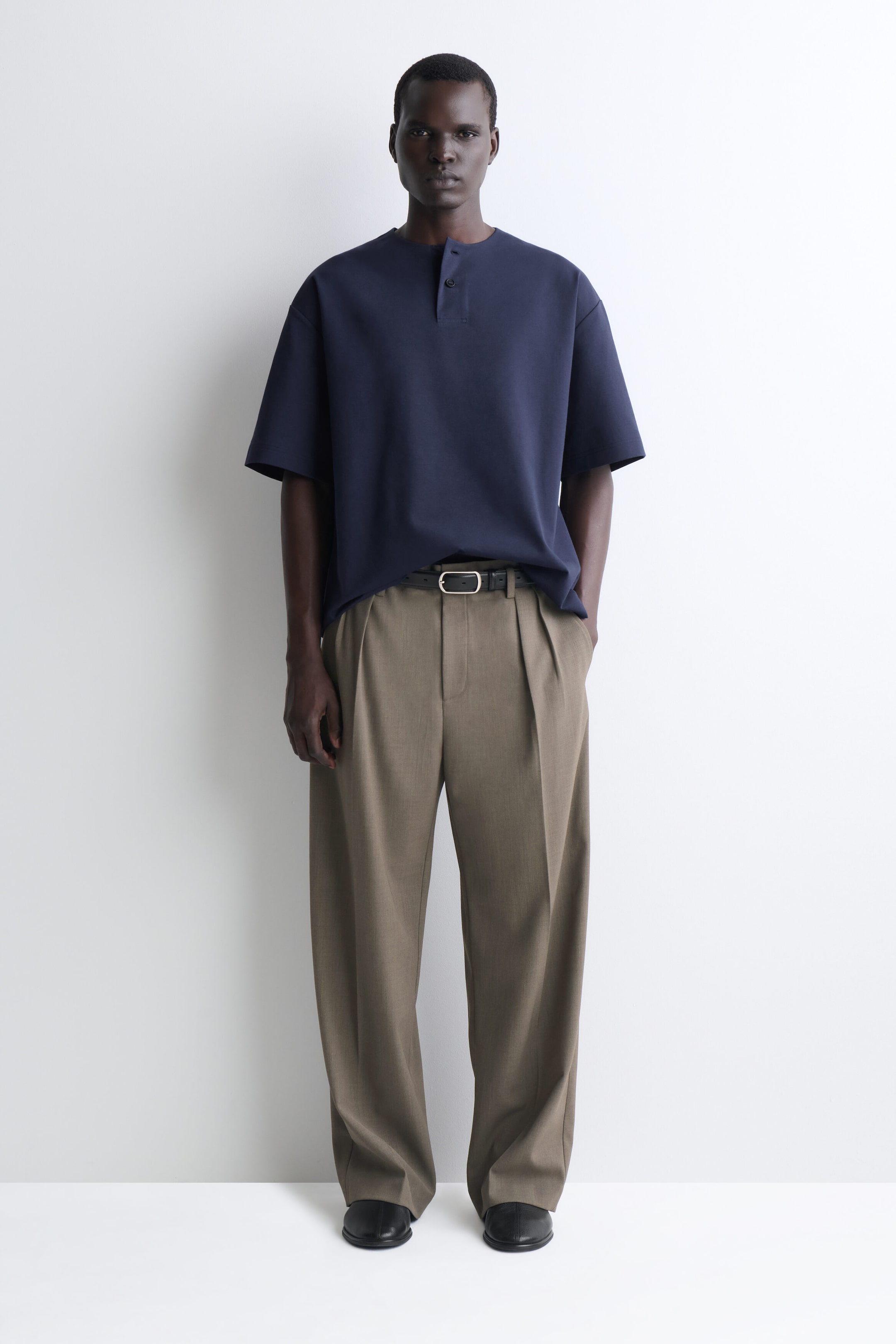 Le t-shirt Henley oversize en jersey de coton est présenté de face, dans une teinte bleu marine, avec des épaules tombantes, des manches courtes amples et une petite patte de boutonnage au col. Le t-shirt tombe souplement et est partiellement rentré dans un pantalon plissé avec une ceinture, mettant en valeur sa coupe décontractée et sa structure souple.
