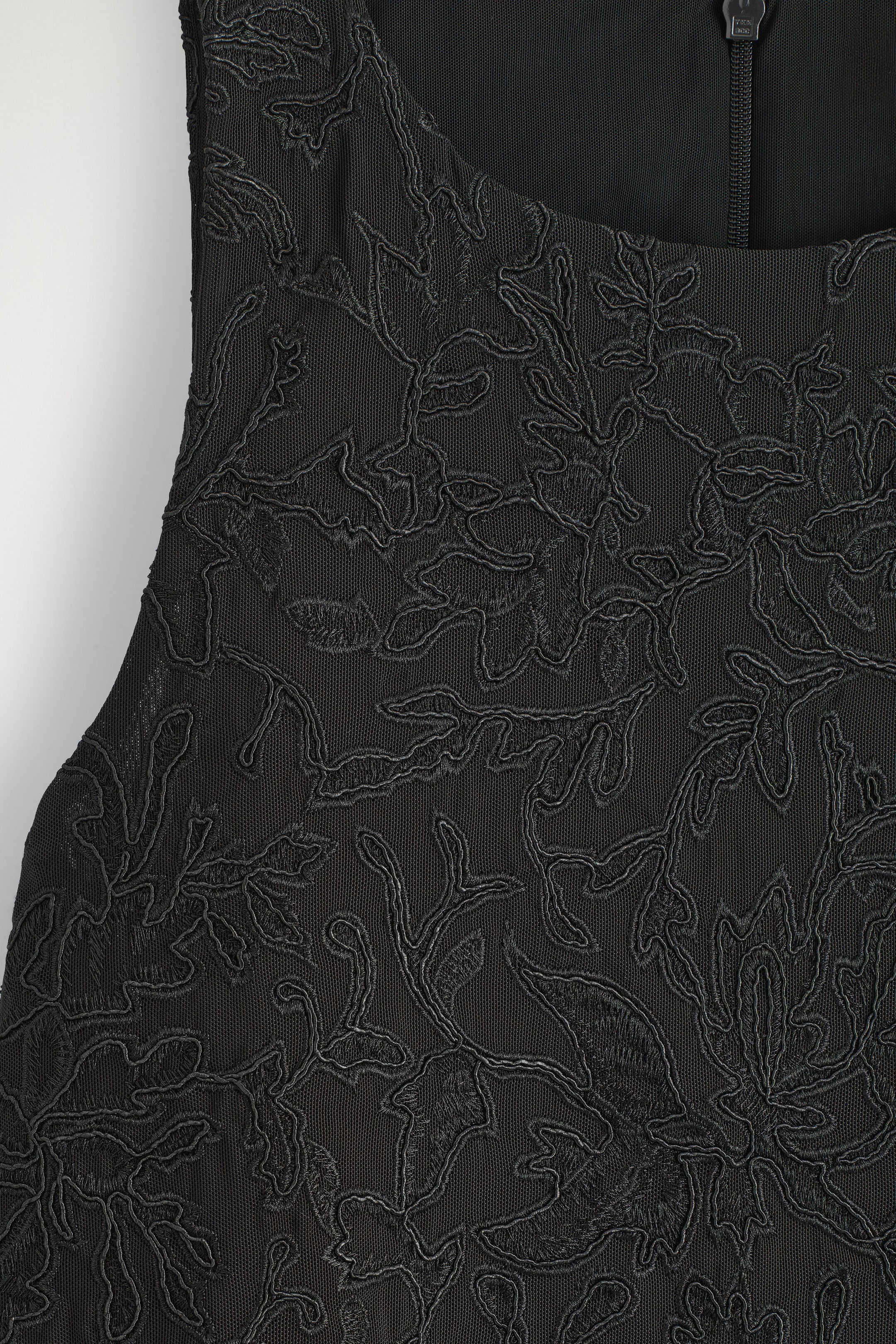 Une prise de vue rapprochée met en valeur la broderie florale noire complexe sur un fond en maille transparente d’un haut. Une fermeture éclair est visible à l’encolure arrondie.