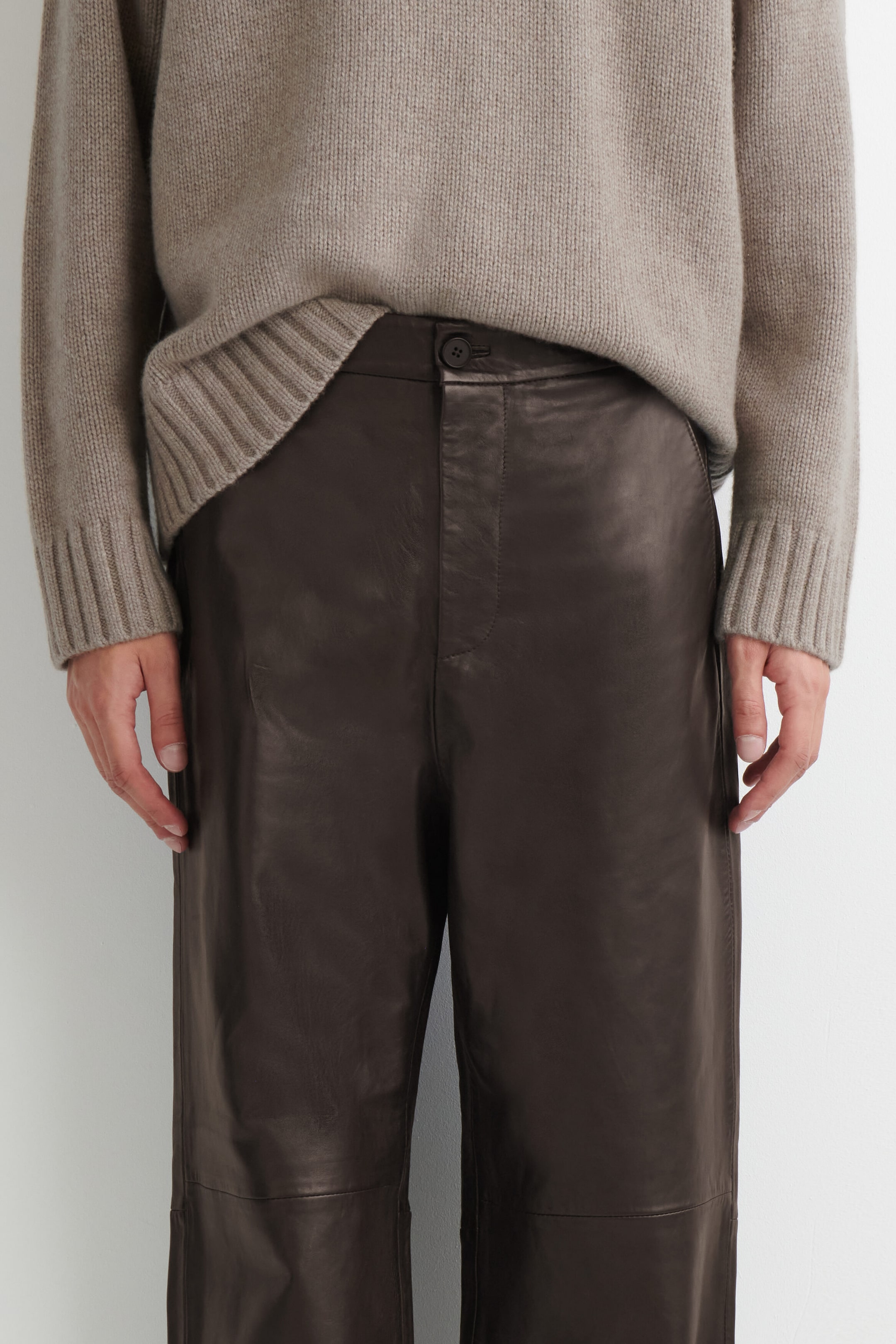 A close-up front view shows the dark brown leather relaxed-fit straight-leg pants, featuring a waistband with a button closure and a smooth leather finish. A beige knit sweater is partially tucked into the front waistband, highlighting the clean design and structure of the pants. A close-up front view shows the dark brown leather relaxed-fit straight-leg pants, featuring a waistband with a button closure and a smooth leather finish. A beige knit sweater is partially tucked into the front waistband, highlighting the clean design and structure of the pants.