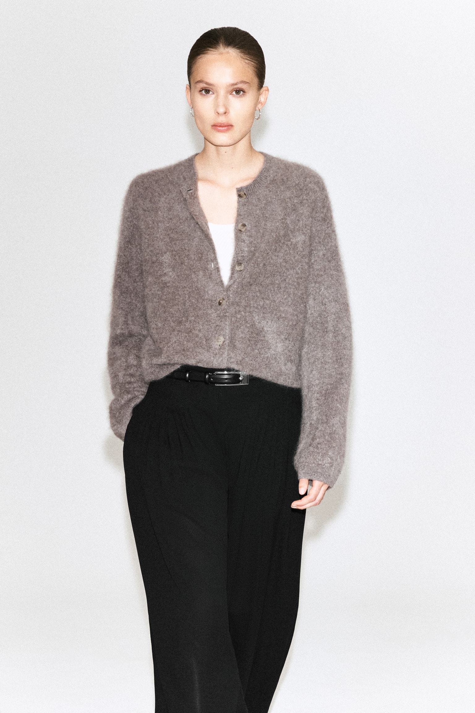Un cardigan en maille duveteuse gris brunâtre présente un col rond et une fermeture boutonnée sur le devant avec plusieurs boutons de couleur claire. Ce cardigan ample, longueur hanches, est porté ouvert sur un haut blanc. - 1