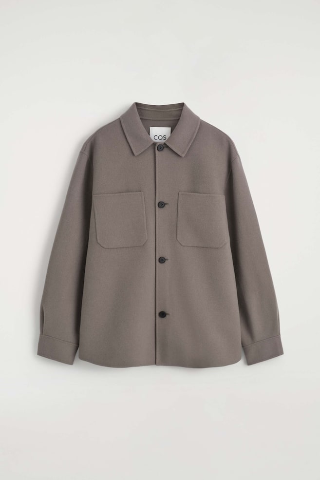 La overshirt in lana double face è presentata in una vista frontale, con un classico colletto a punta, chiusura frontale a quattro bottoni e due ampie tasche applicate simmetriche sul petto. Il capo mostra maniche lunghe con polsini strutturati e un orlo dritto in un colore taupe-grigio tinta unita. - 1