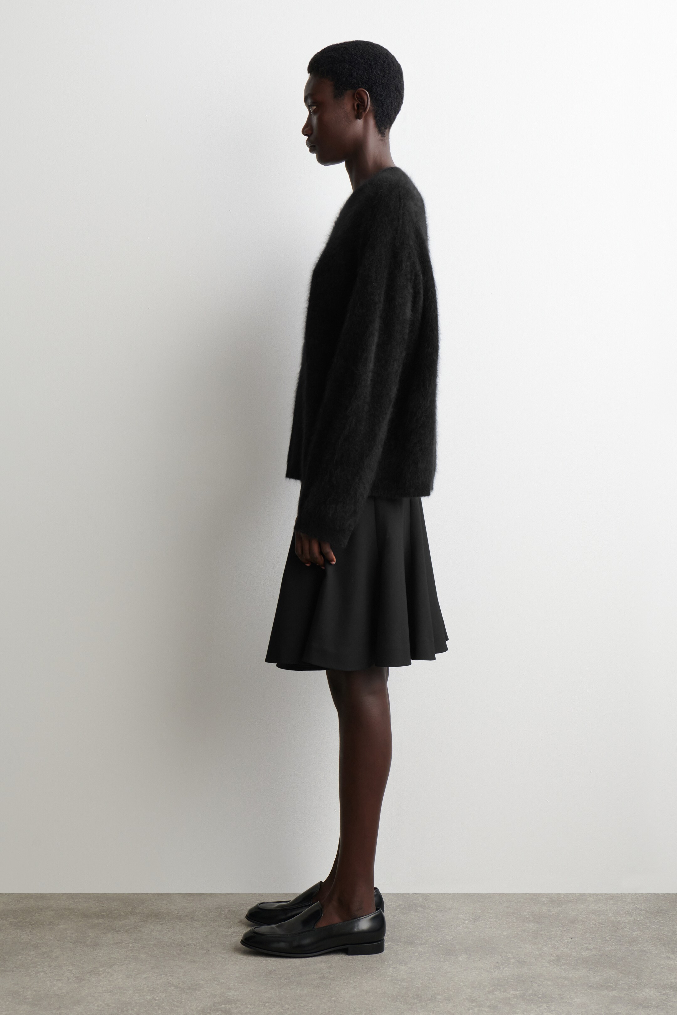 Se større billede: En sort, fuzzy strikket sweater med afslappet pasform og lange ærmer styles med en sort, udsvinget mininederdel. Ensemblet fuldendes med sorte læderloafers, alt præsenteret fra en sideprofil.
