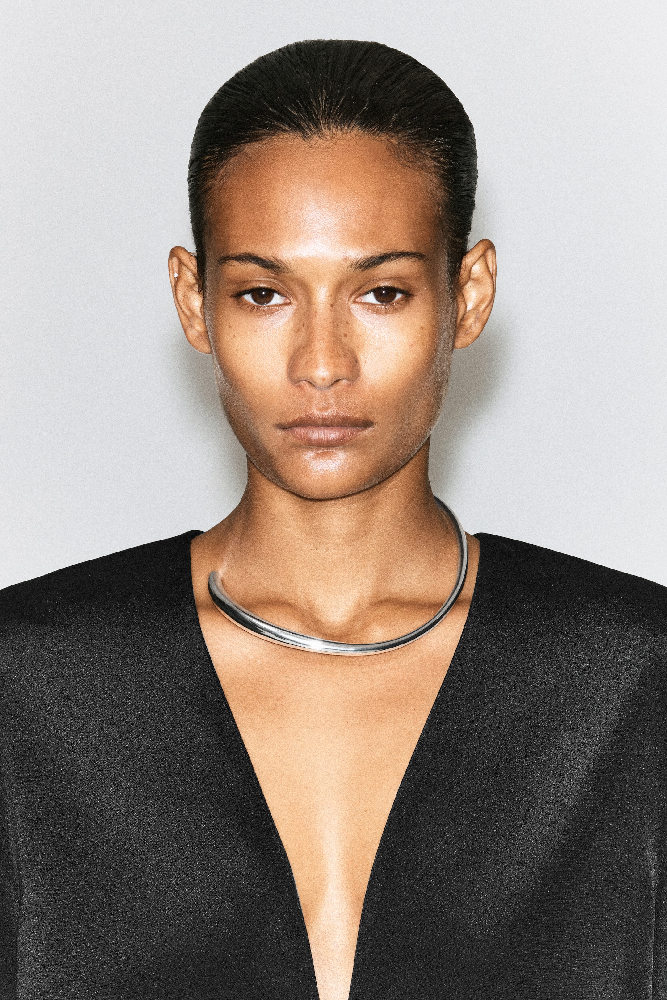 Un collier ras du cou en ton argenté poli, au design épais et tubulaire, est porté par le mannequin et repose à la base du cou. Cette pièce de joaillerie minimaliste est stylisée avec un vêtement noir doté d’un profond décolleté en V. Un collier ras du cou en ton argenté poli, au design épais et tubulaire, est porté par le mannequin et repose à la base du cou. Cette pièce de joaillerie minimaliste est stylisée avec un vêtement noir doté d’un profond décolleté en V.