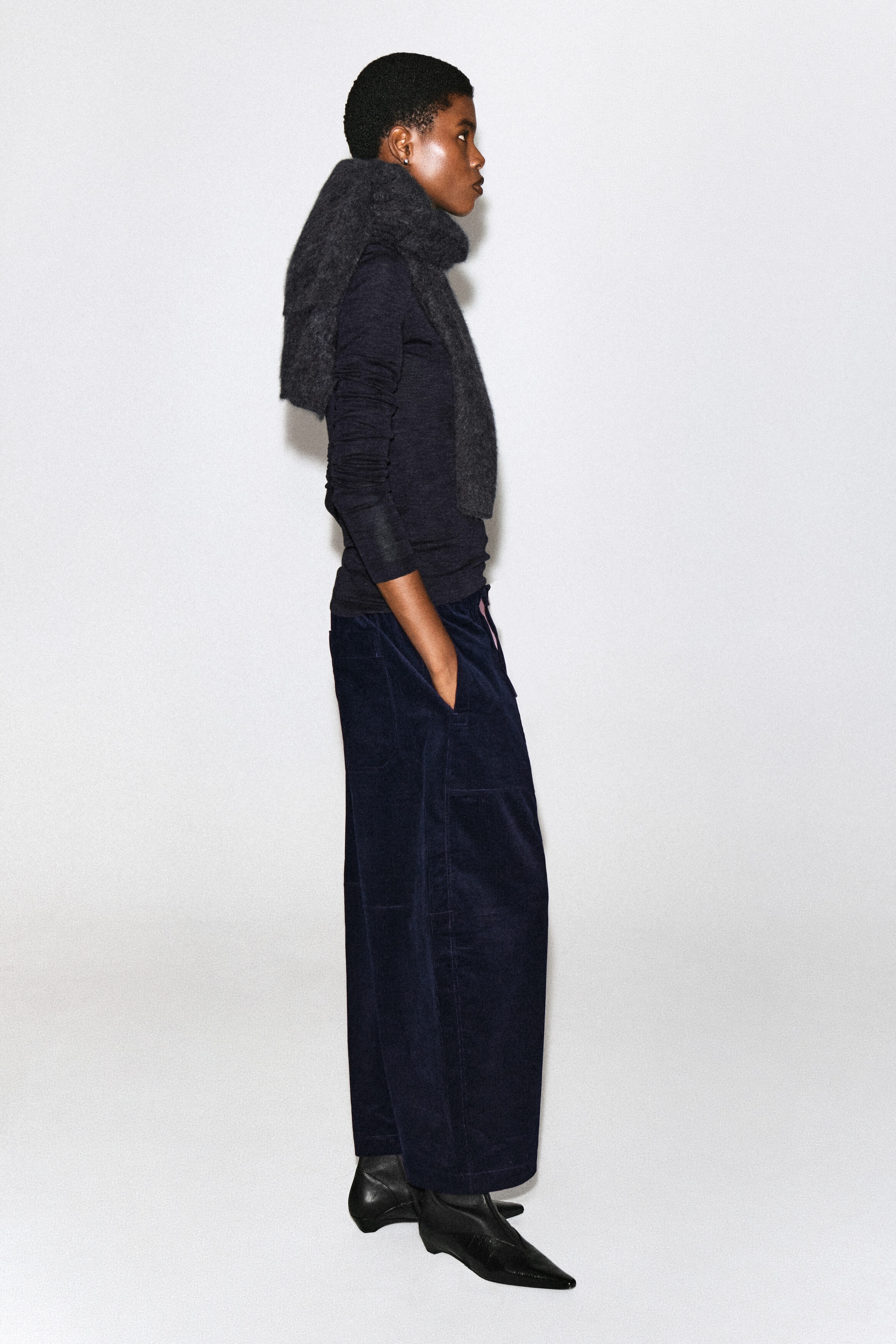 Corduroy barrel-leg pants in dark navy blue are worn by the model, who is standing in profile with one hand in the side pocket. The pants are styled with a dark long-sleeve top, a gray knit scarf draped over the shoulders, and black pointed-toe boots. Corduroy barrel-leg pants in dark navy blue are worn by the model, who is standing in profile with one hand in the side pocket. The pants are styled with a dark long-sleeve top, a gray knit scarf draped over the shoulders, and black pointed-toe boots.