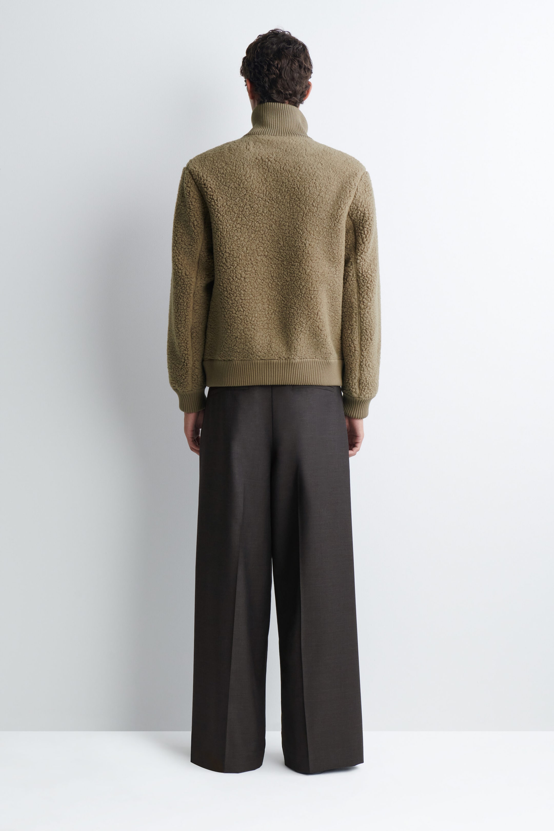 An olive-green textured fleece jacket is being worn by the model, shown from the back. The jacket features a high ribbed collar, ribbed cuffs, and a ribbed hem, and is styled with dark brown wide-leg trousers. An olive-green textured fleece jacket is being worn by the model, shown from the back. The jacket features a high ribbed collar, ribbed cuffs, and a ribbed hem, and is styled with dark brown wide-leg trousers.