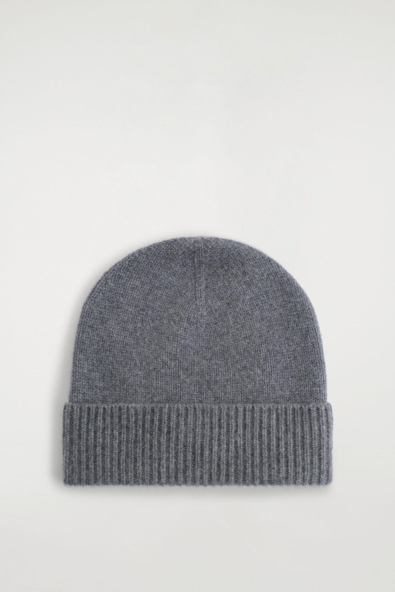 A grey cashmere beanie is resting in a front view, showcasing a soft fine-knit rounded crown and a thick, vertically ribbed turn-up cuff.