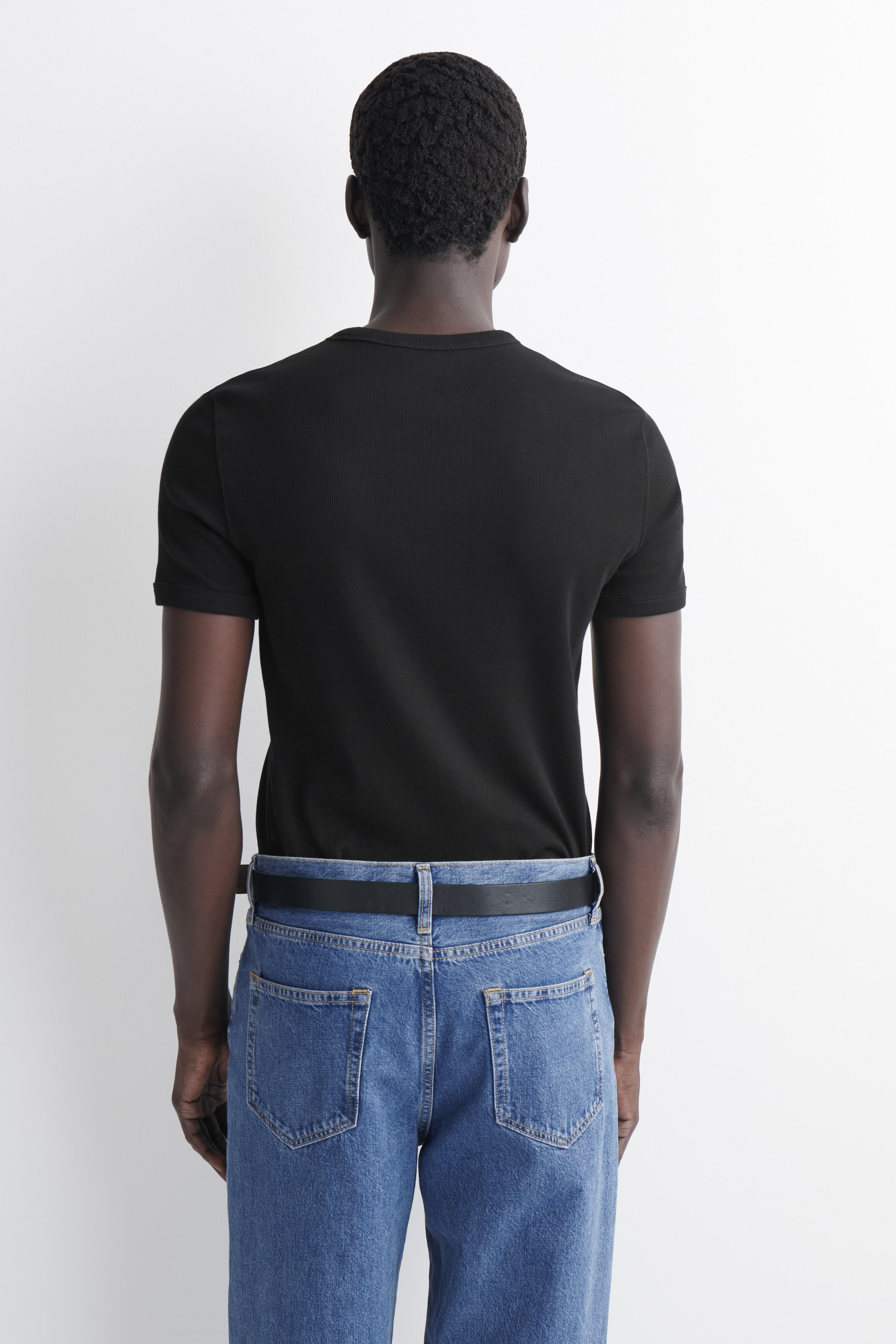 Grotere afbeelding bekijken: Het zwarte geribbelde T-shirt met korte mouwen wordt van achteren getoond, met een aansluitende pasvorm, een ronde hals en korte mouwen die de armen omsluiten, en het is ingestopt in een jeans.