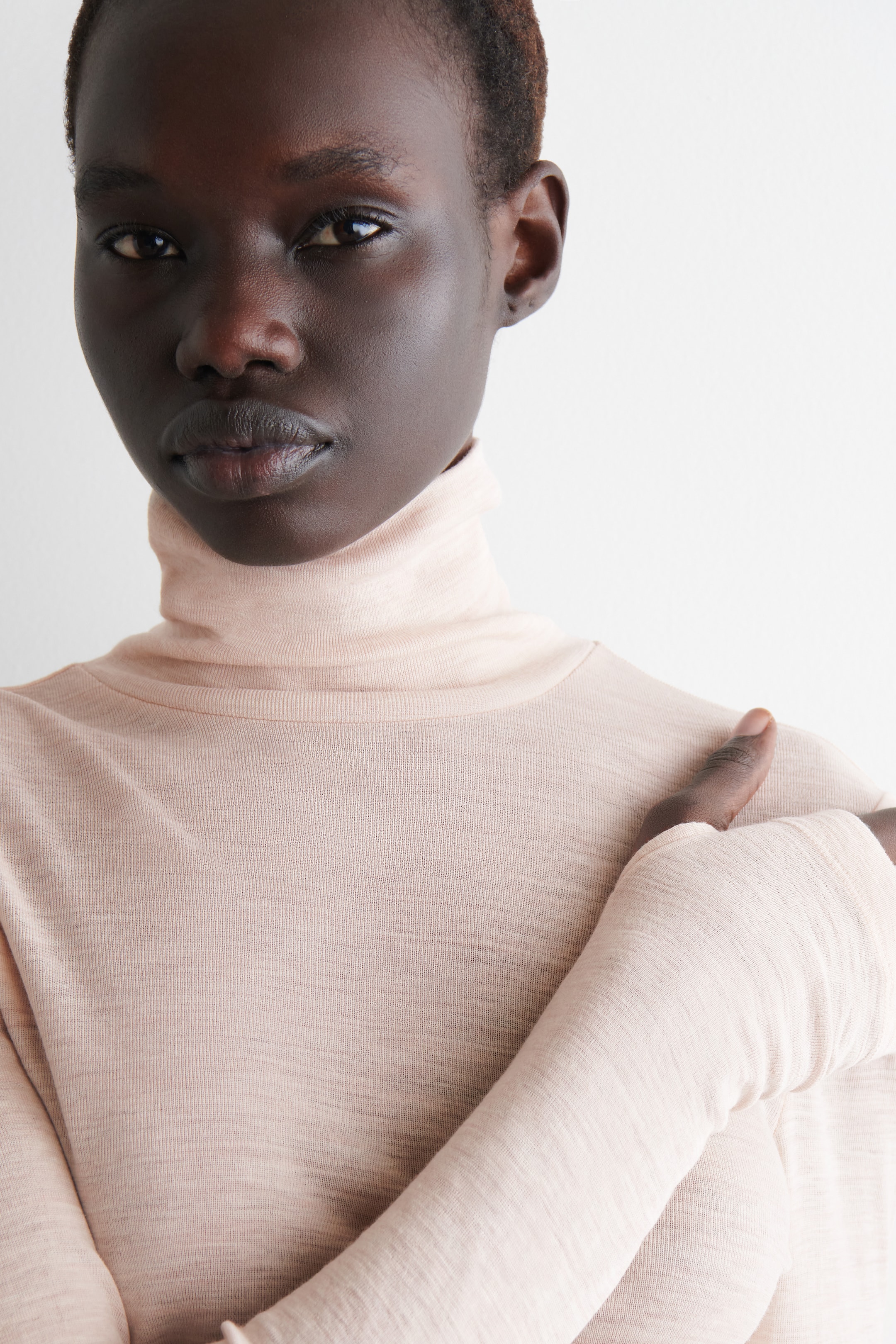 View larger image: A pale peach turtleneck top is being worn, featuring a close-fitting collar and long sleeves. The lightweight knit fabric is showing subtle horizontal texture, and a hand is resting on the shoulder, showcasing the sleeve's form-fitting design.