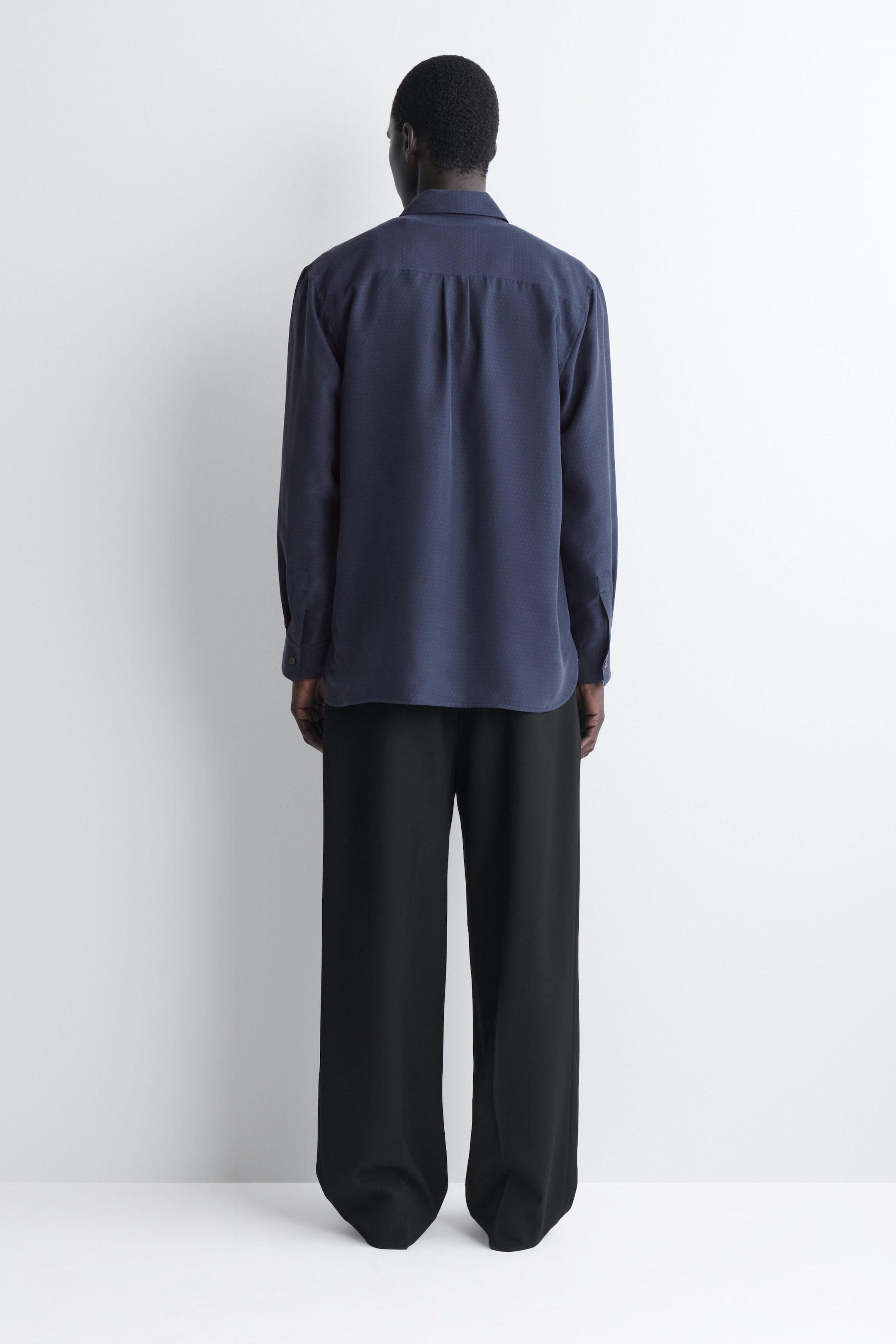 A sandwashed silk shirt in navy blue is shown from the back, featuring a relaxed fit, pointed collar, and pleated back yoke. The long-sleeve shirt drapes loosely and is styled with wide-leg black trousers. A sandwashed silk shirt in navy blue is shown from the back, featuring a relaxed fit, pointed collar, and pleated back yoke. The long-sleeve shirt drapes loosely and is styled with wide-leg black trousers.