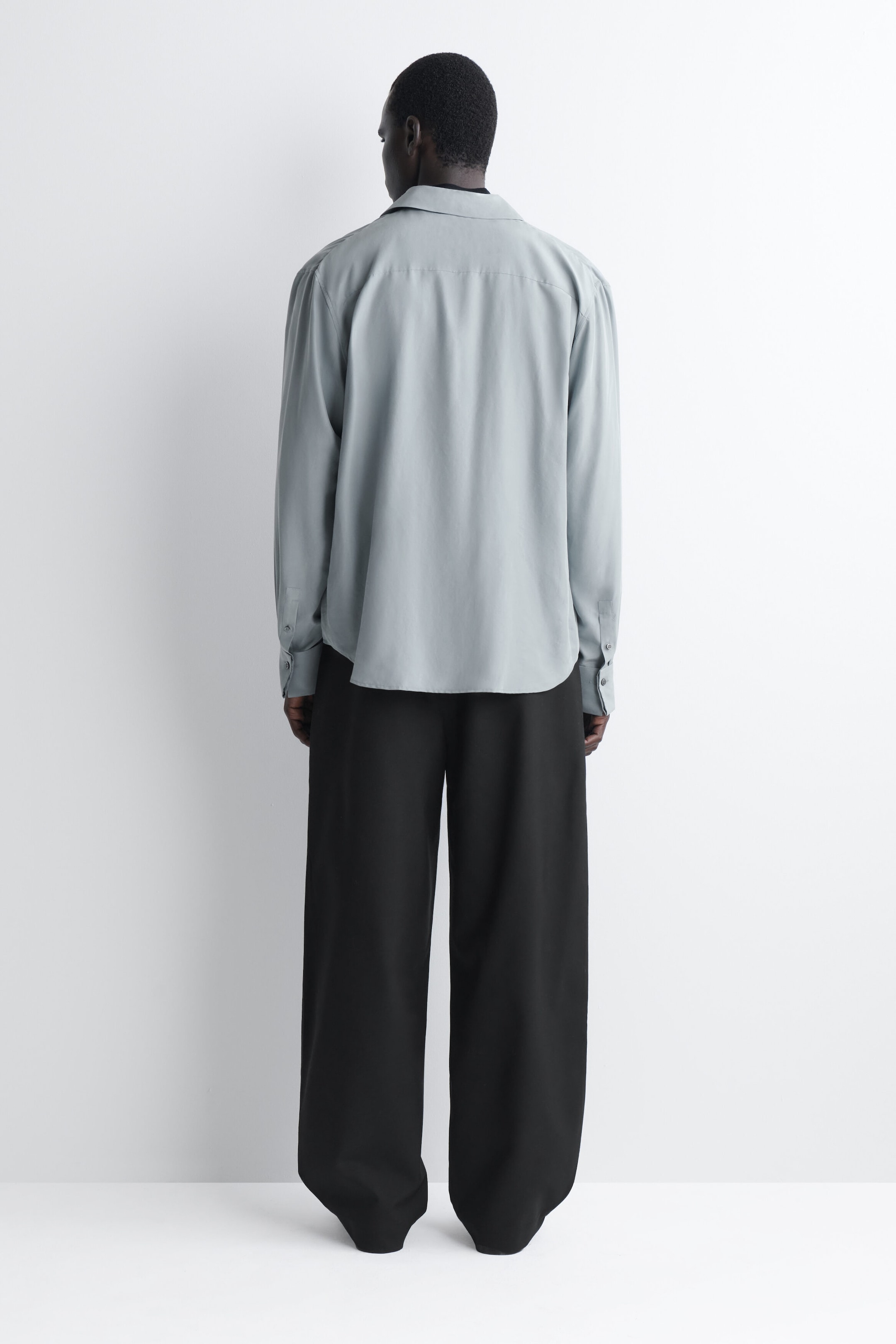 Die schwarzen Barrel-Leg-Hosen im Relaxed Fit werden von hinten gezeigt und betonen ihre weite, dezent geschwungene Silhouette sowie den fließenden Fall. Ein hellgraues Langarmshirt wird locker über dem Bund getragen und teilweise über den oberen Bereich der Hose gelegt. Die schwarzen Barrel-Leg-Hosen im Relaxed Fit werden von hinten gezeigt und betonen ihre weite, dezent geschwungene Silhouette sowie den fließenden Fall. Ein hellgraues Langarmshirt wird locker über dem Bund getragen und teilweise über den oberen Bereich der Hose gelegt.