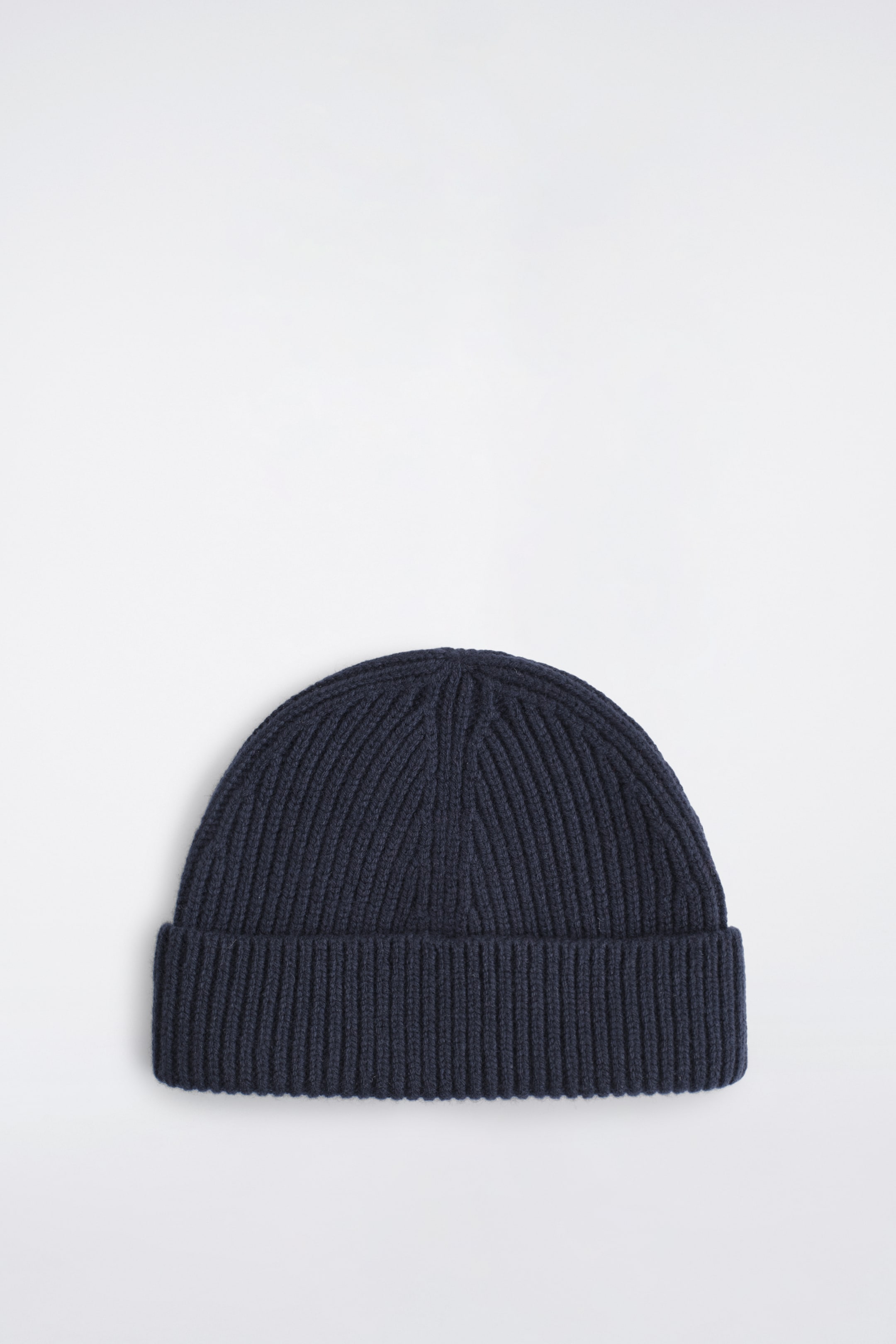 Se større billede: En kraftig ribstrikket uldbeanie i mørk marineblå vender forfra og fremhæver sin tykke ribstrikkede struktur og en bred opfoldet kant. Beanie’en viser en rundet top med synlige strikkede indtagningslinjer, der smalner ind mod midten i toppen.