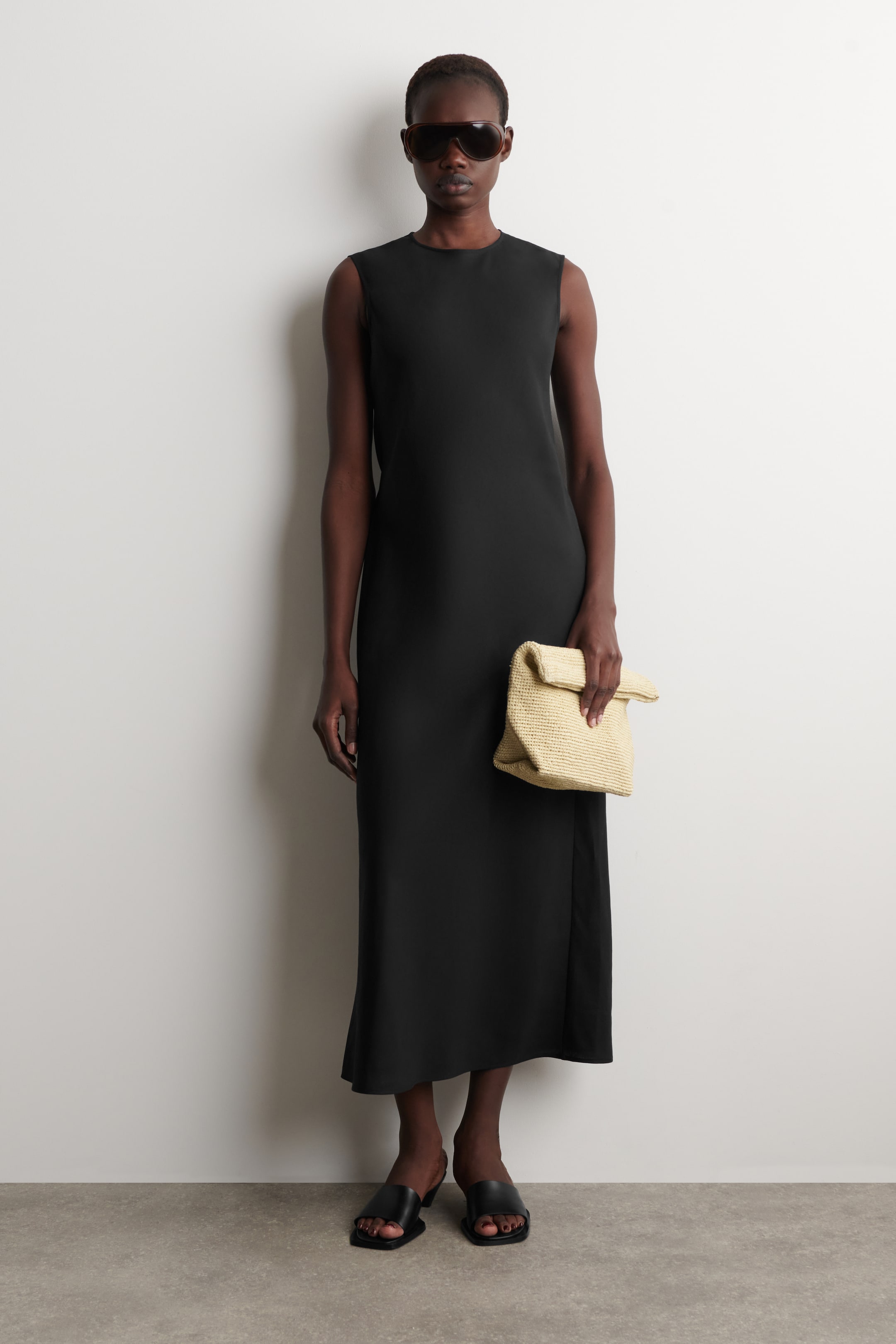 הצגת תמונה גדולה יותר: השמלה השחורה מציגה עיצוב ללא שרוולים עם צווארון עגול מזווית קדמית, המדגישה את הצללית הישרה שלה. היא מגיעה לאורך הקרסול ומציגה חריץ גבוה בצד הימני של הלובש.
