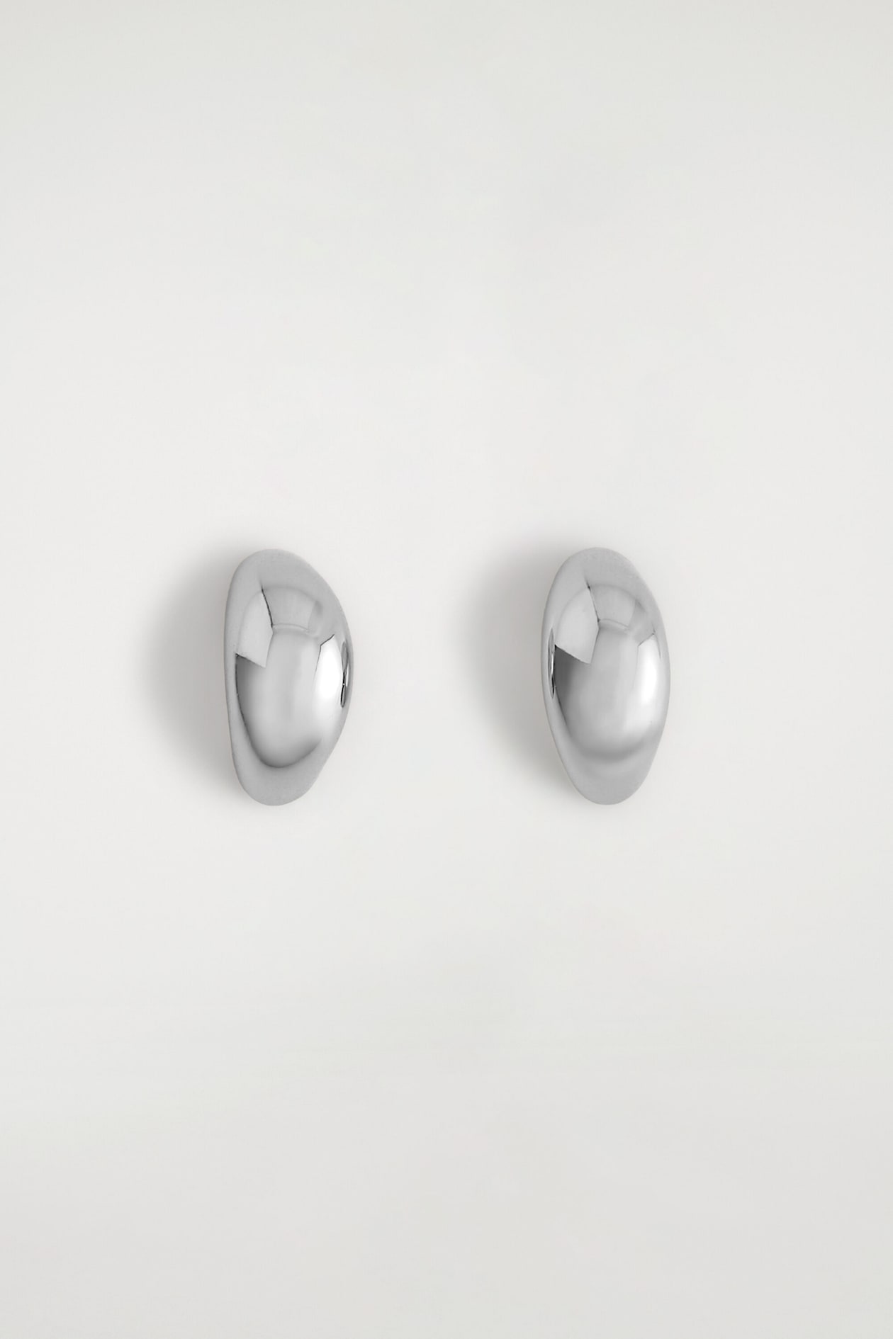 Une paire de boucles d’oreilles demi-créoles sculpturales est présentée de face, affichant une finition argentée très polie. Les boucles d’oreilles présentent une forme organique lisse, bombée et arrondie.