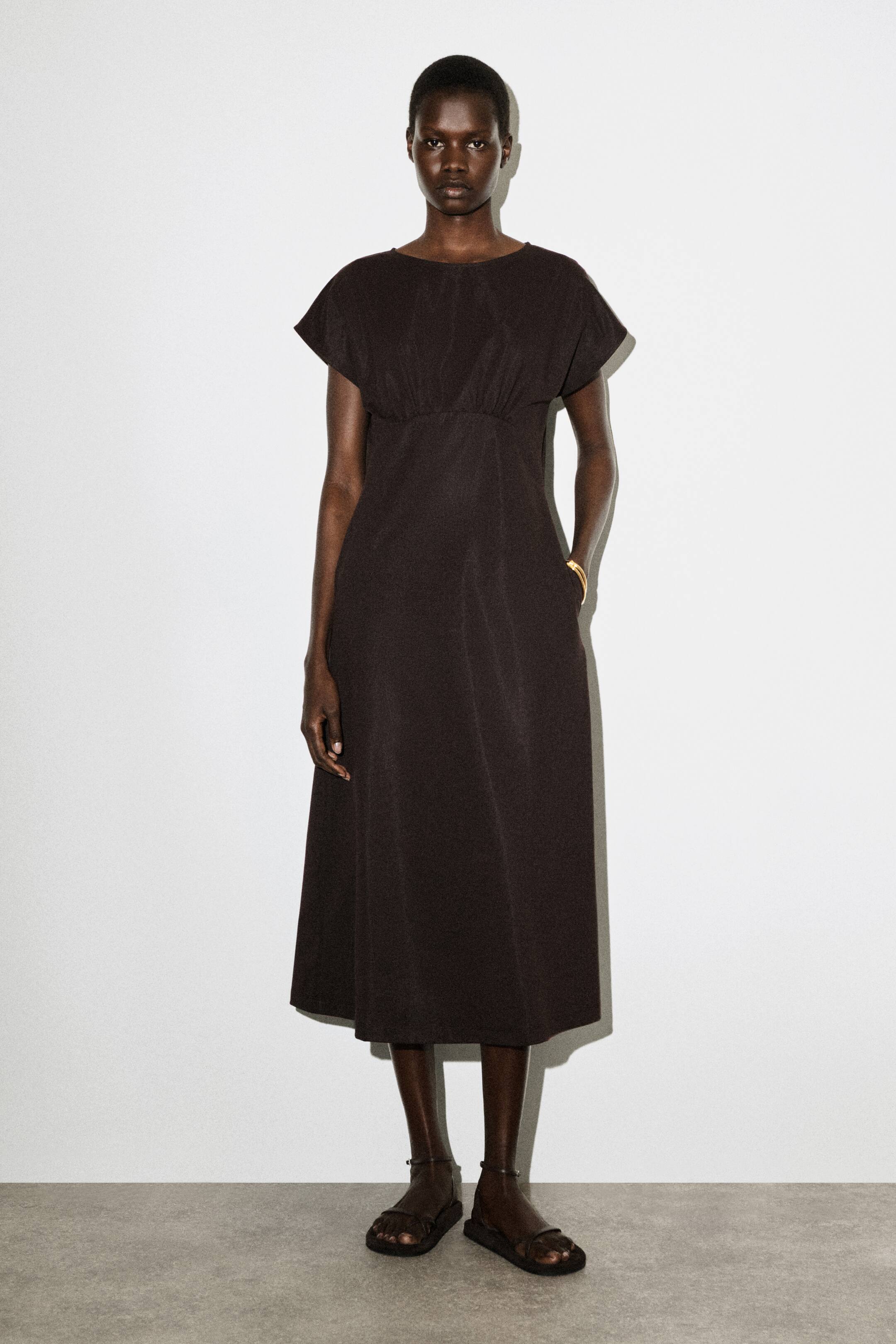 Agrandir l'image: Une robe midi marron foncé présente un col rond et des manches courtes et larges de type "cap". Elle se distingue par un corsage froncé au-dessus d'une taille empire, s'épanouissant en une jupe évasée en ligne A avec une poche latérale visible.