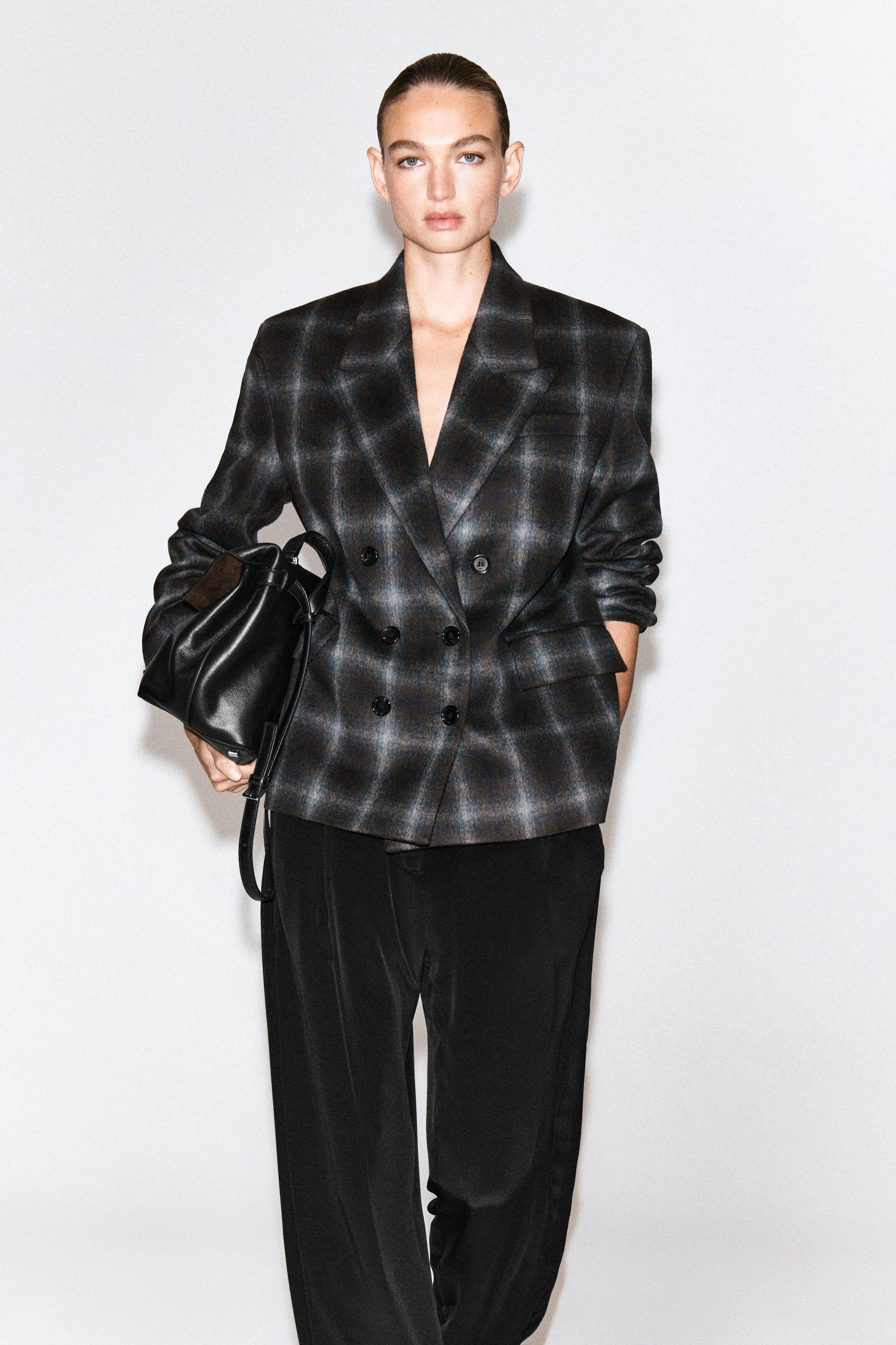 Un blazer croisé en mélange de laine à motif à carreaux foncé est porté par le mannequin. Ce blazer, doté de revers à encoche et de fermetures à boutons, est associé à un pantalon noir, tandis que le mannequin tient un sac en cuir noir sous un bras.