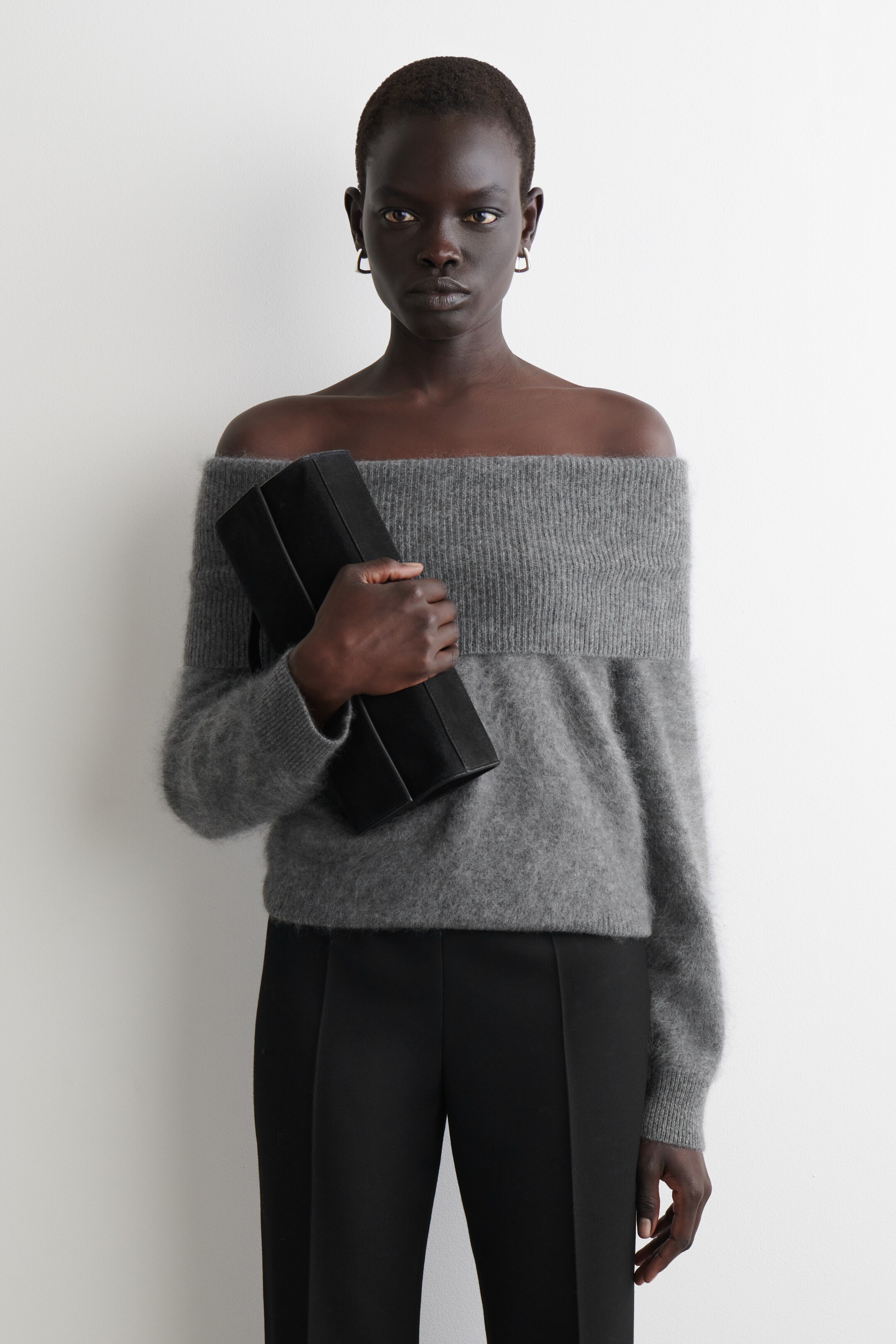 Ingrandisci l'immagine: Un maglione grigio a spalle scoperte presenta una banda larga a coste sulla linea delle spalle e una trama morbida e pelosa, che si estende fino ai fianchi. È abbinato a pantaloni neri che presentano una cucitura verticale sul davanti, e una pochette nera strutturata viene tenuta in mano.