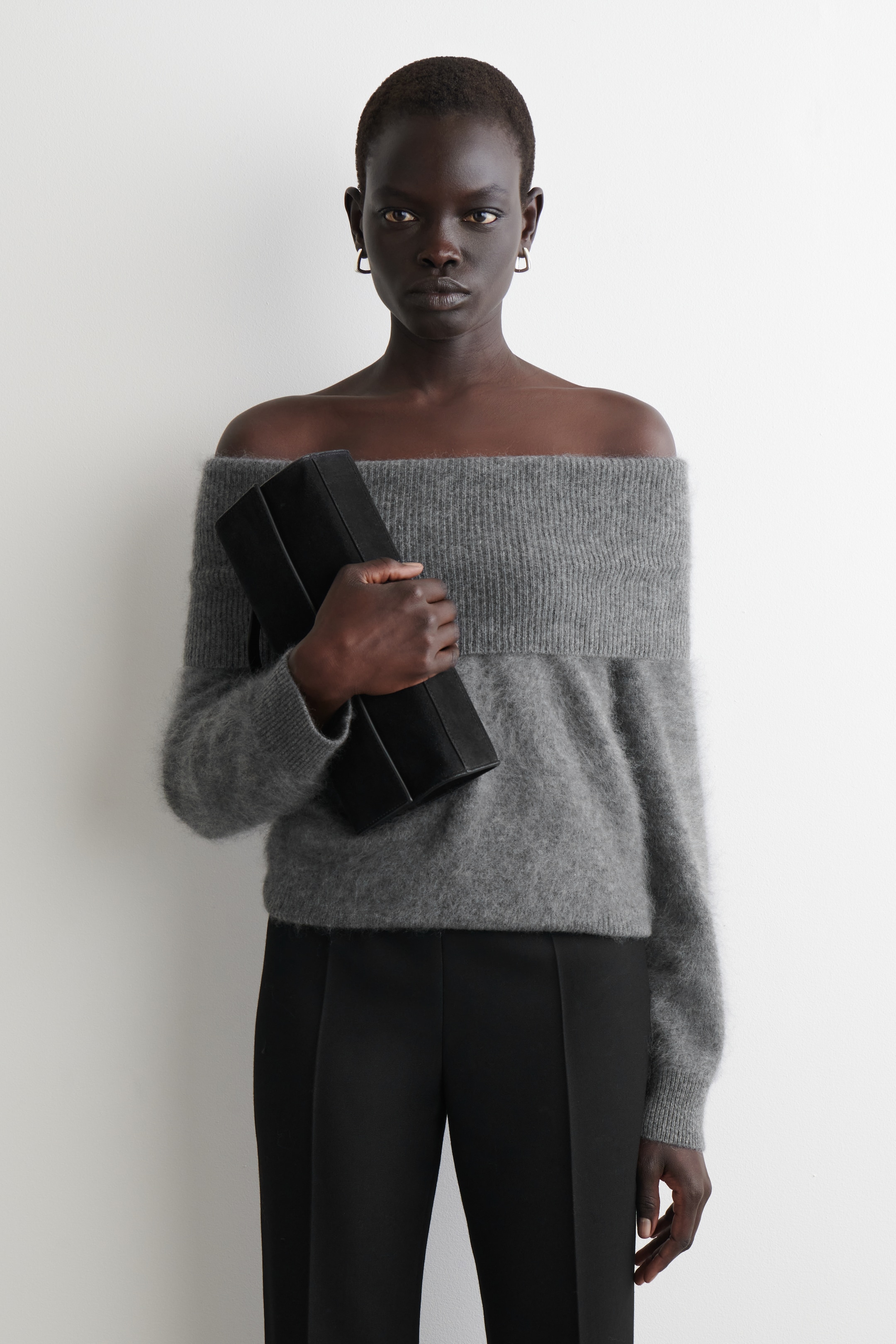 Ver imagen más grande: Un suéter gris sin hombros presenta una banda ancha acanalada en la línea de los hombros y una textura suave y esponjosa, extendiéndose hasta las caderas. Se combina con unos pantalones negros que presentan una costura frontal vertical, y se sostiene un clutch estructurado negro.