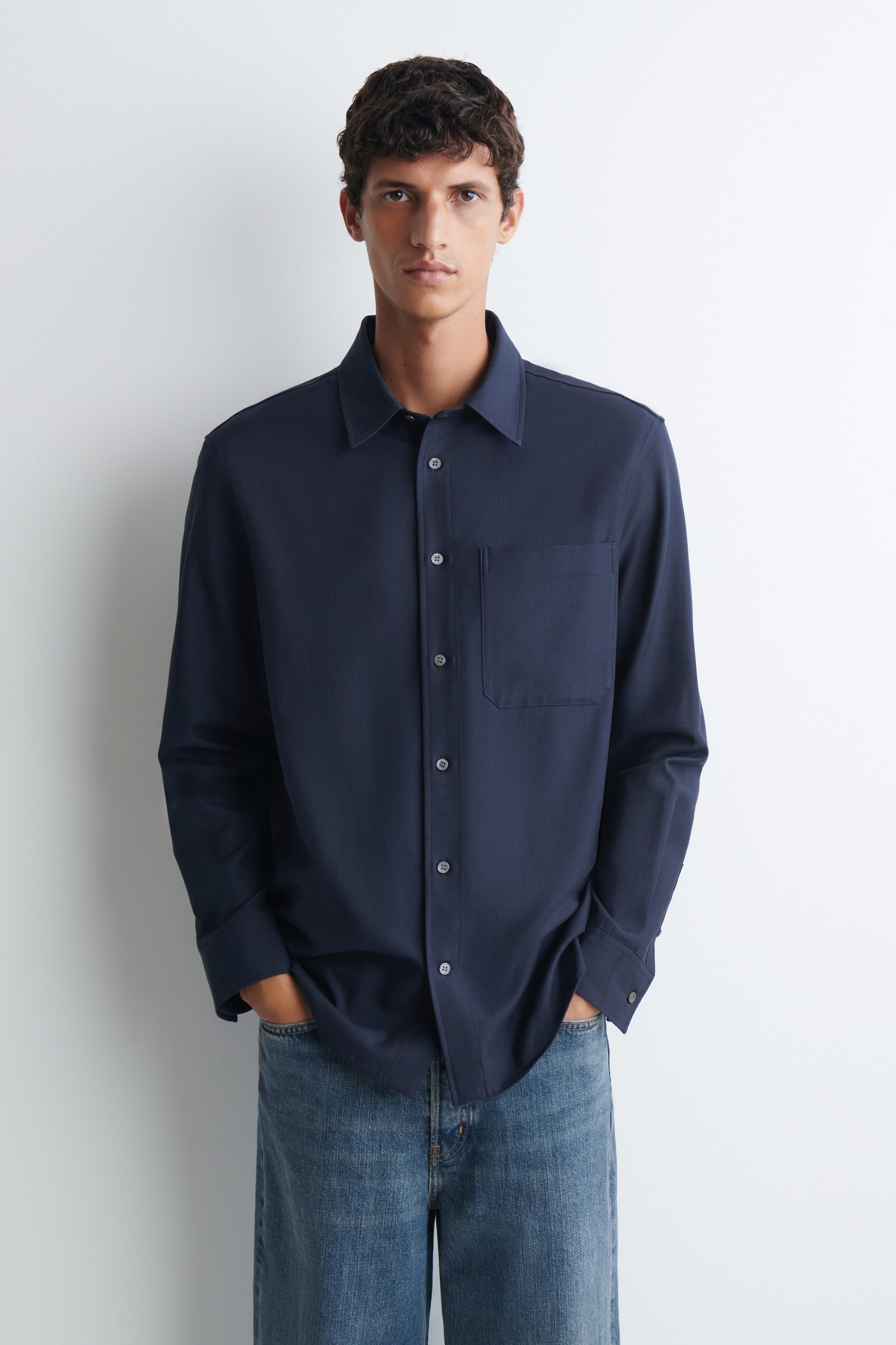 A navy blue relaxed twill shirt featuring a pointed collar, button-up front, and a single chest pocket is being worn by the model. The model is pairing the long-sleeved shirt with blue jeans, resting hands inside the trouser pockets.