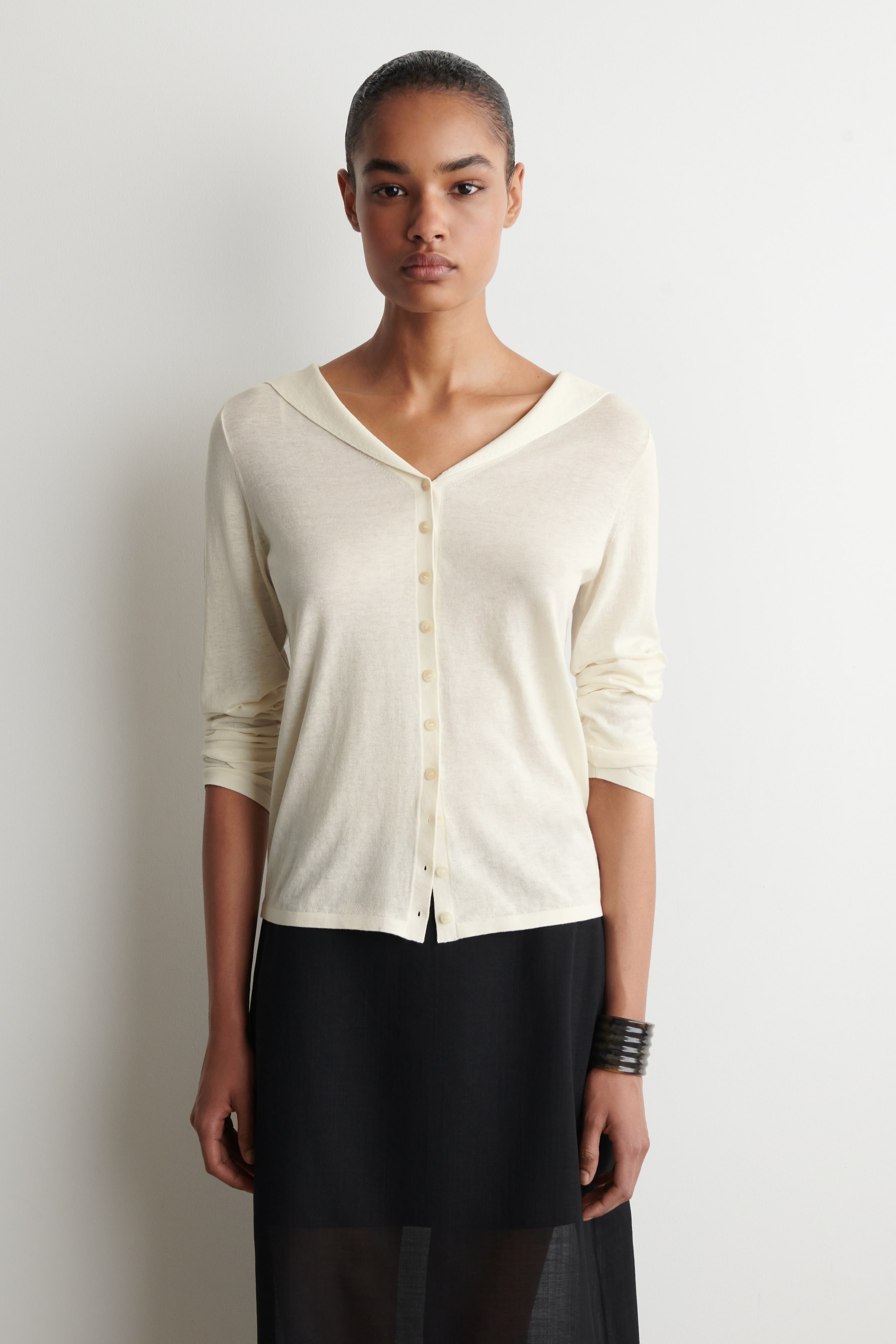 Visualizza l'immagine ingrandita: Un cardigan in maglia leggera color crema presenta uno scollo a V con un ampio colletto a scialle. È dotato di una chiusura frontale completa con piccoli bottoni di colore chiaro, e le sue maniche lunghe sono spinte fino a una lunghezza tre quarti.
