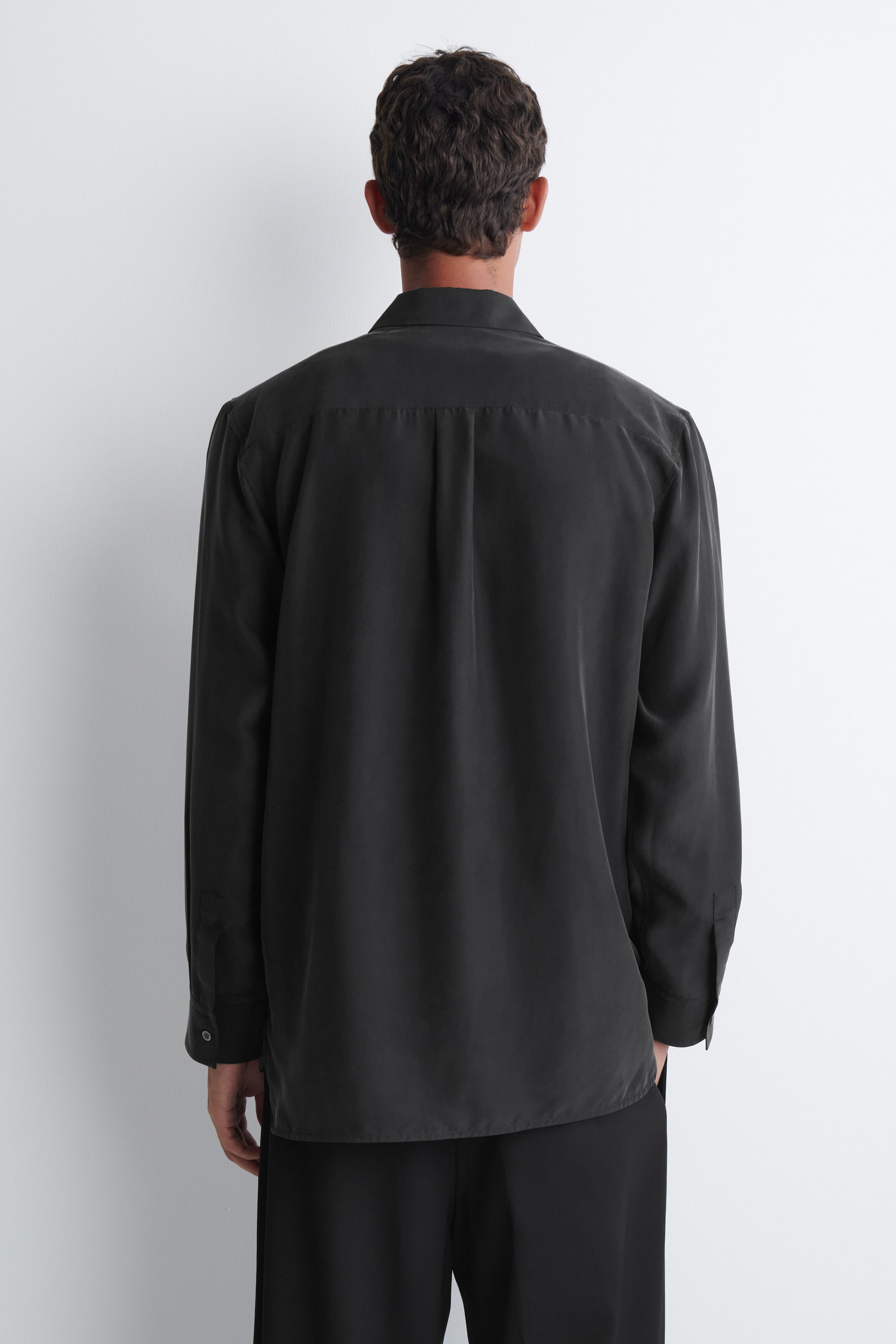 The model is wearing a black sandwashed silk shirt, shown from the back. The long-sleeved shirt features a relaxed fit, a classic collar, and a back yoke with a central box pleat. The model is wearing a black sandwashed silk shirt, shown from the back. The long-sleeved shirt features a relaxed fit, a classic collar, and a back yoke with a central box pleat.