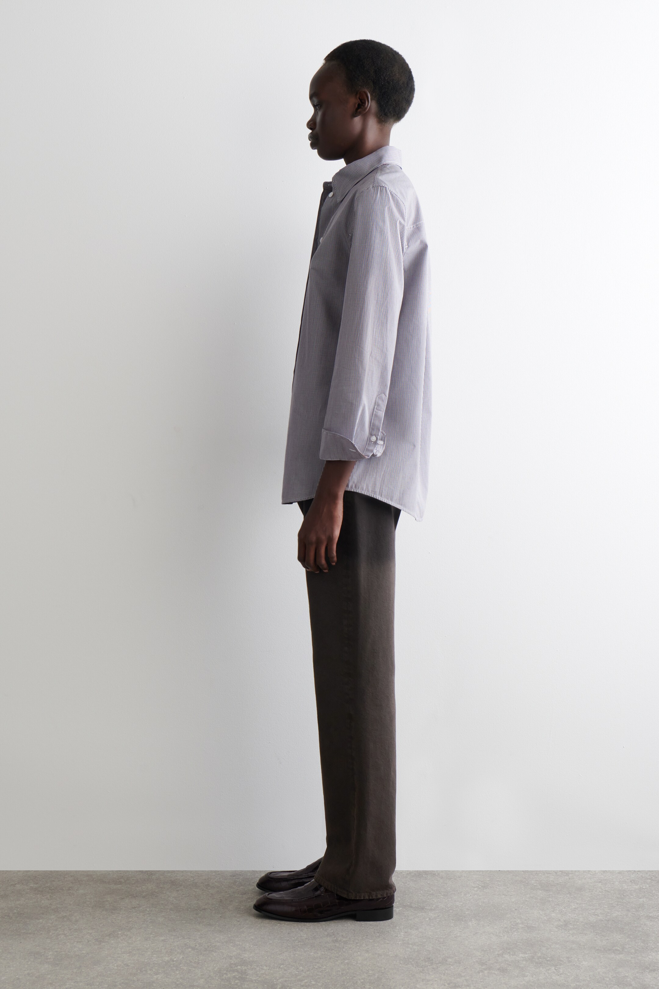 Agrandir l'image: Une chemise gris clair à boutons, semblant ample avec les manches retroussées jusqu'aux avant-bras, est portée avec un pantalon large marron foncé et des mocassins texturés marron foncé. La vue de profil met en évidence le drapé décontracté de la chemise et la coupe droite du pantalon.