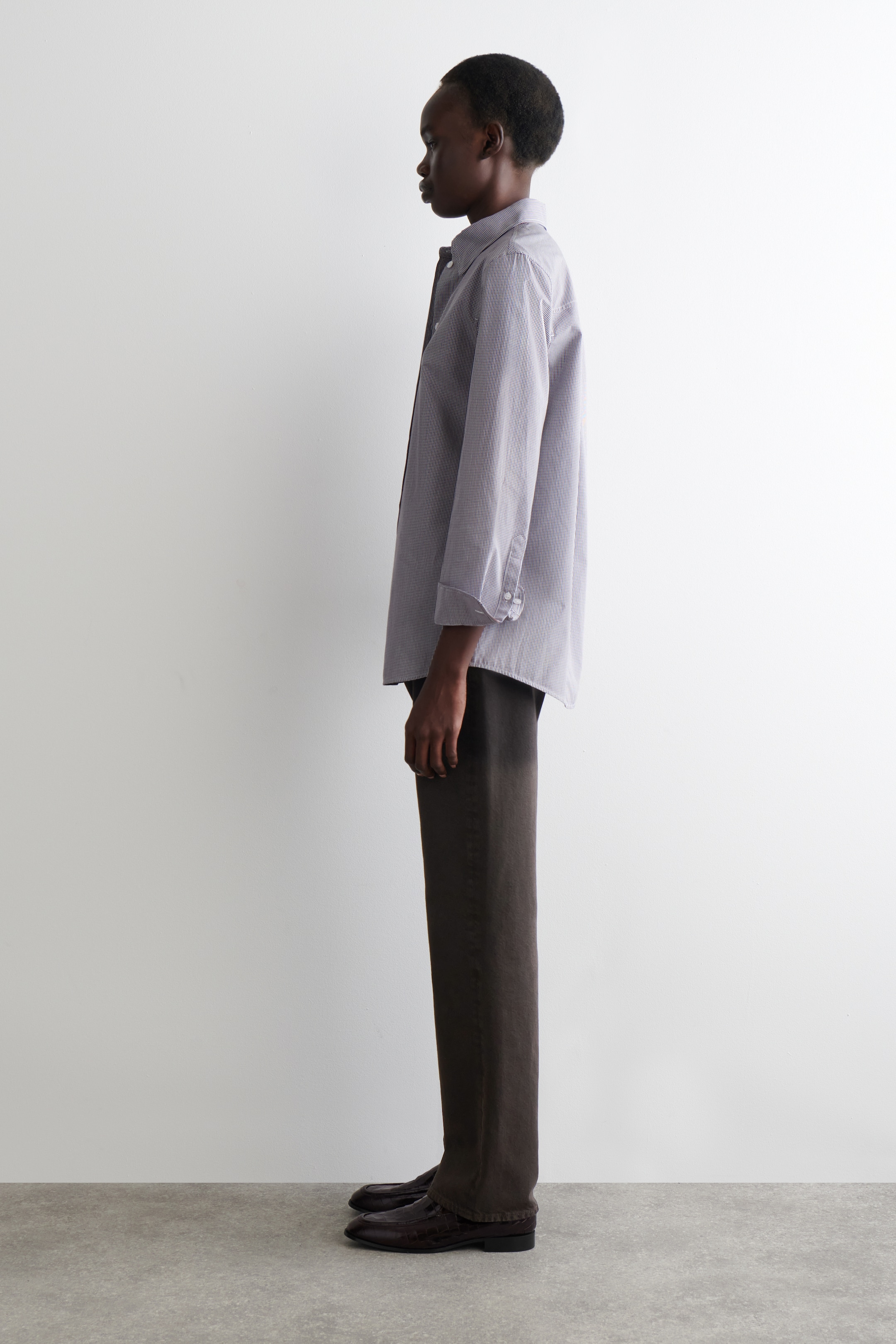 Visualizza immagine più grande: Una camicia grigio chiaro con bottoni, che appare ampia con le maniche arrotolate fino agli avambracci, viene indossata con pantaloni a gamba larga marrone scuro e mocassini marrone scuro con texture. La vista di profilo laterale mette in evidenza il drappeggio rilassato della camicia e il taglio dritto dei pantaloni.