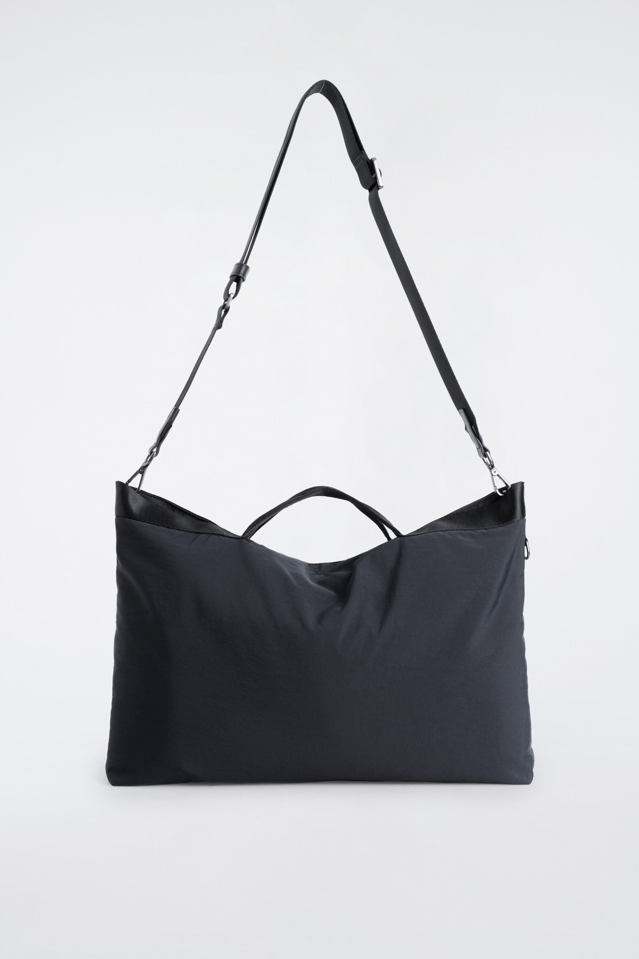 A black fabric bag is shown from the front, featuring a slouchy rectangular shape with dual top handles and a long adjustable shoulder strap attached with metal clasps.